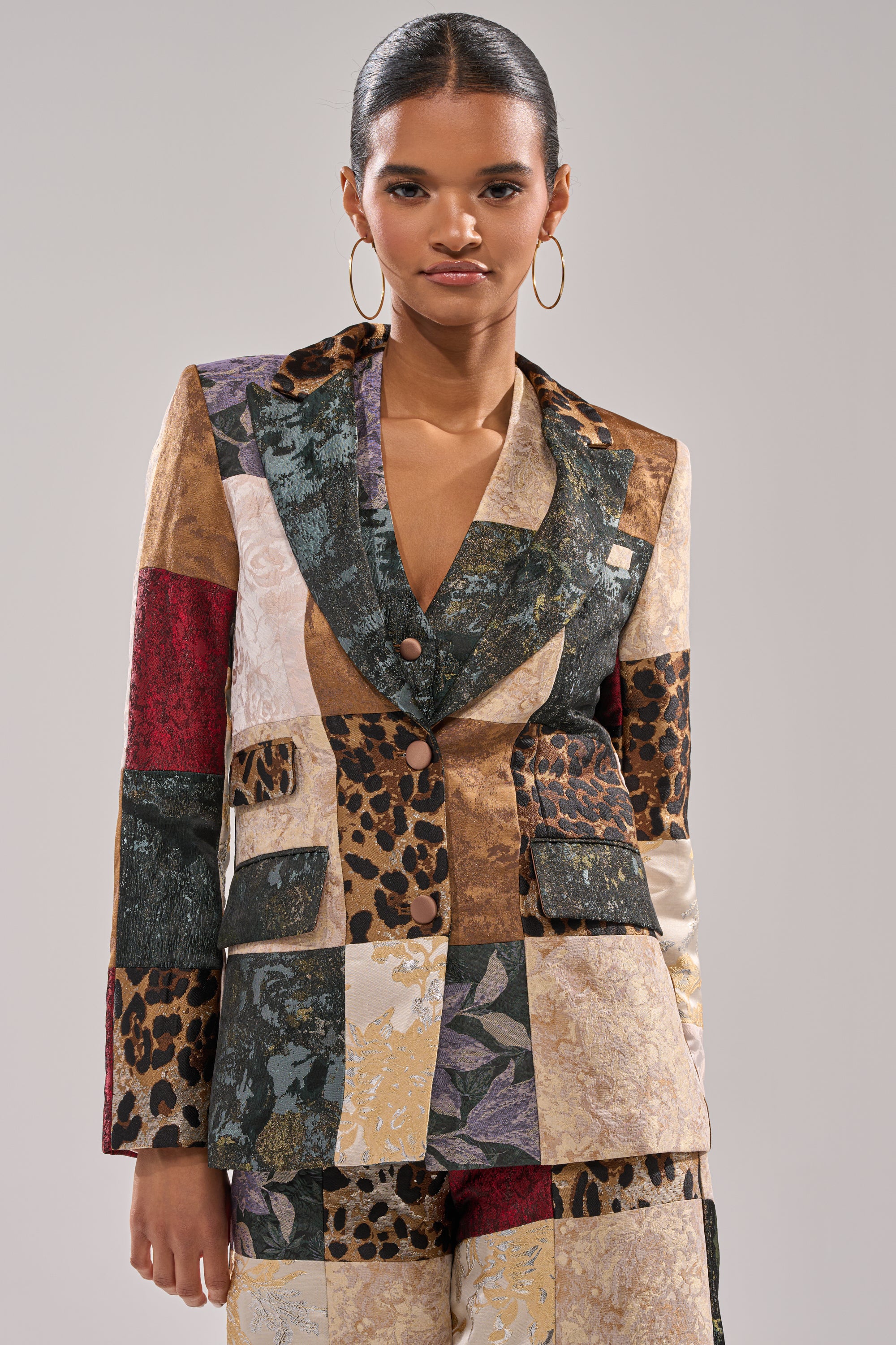 A woman with slicked-back hair wears large hoop earrings and the MADE YOU LOOK PATCHWORK BLAZER, featuring animal prints and florals, paired with matching tailored trousers.