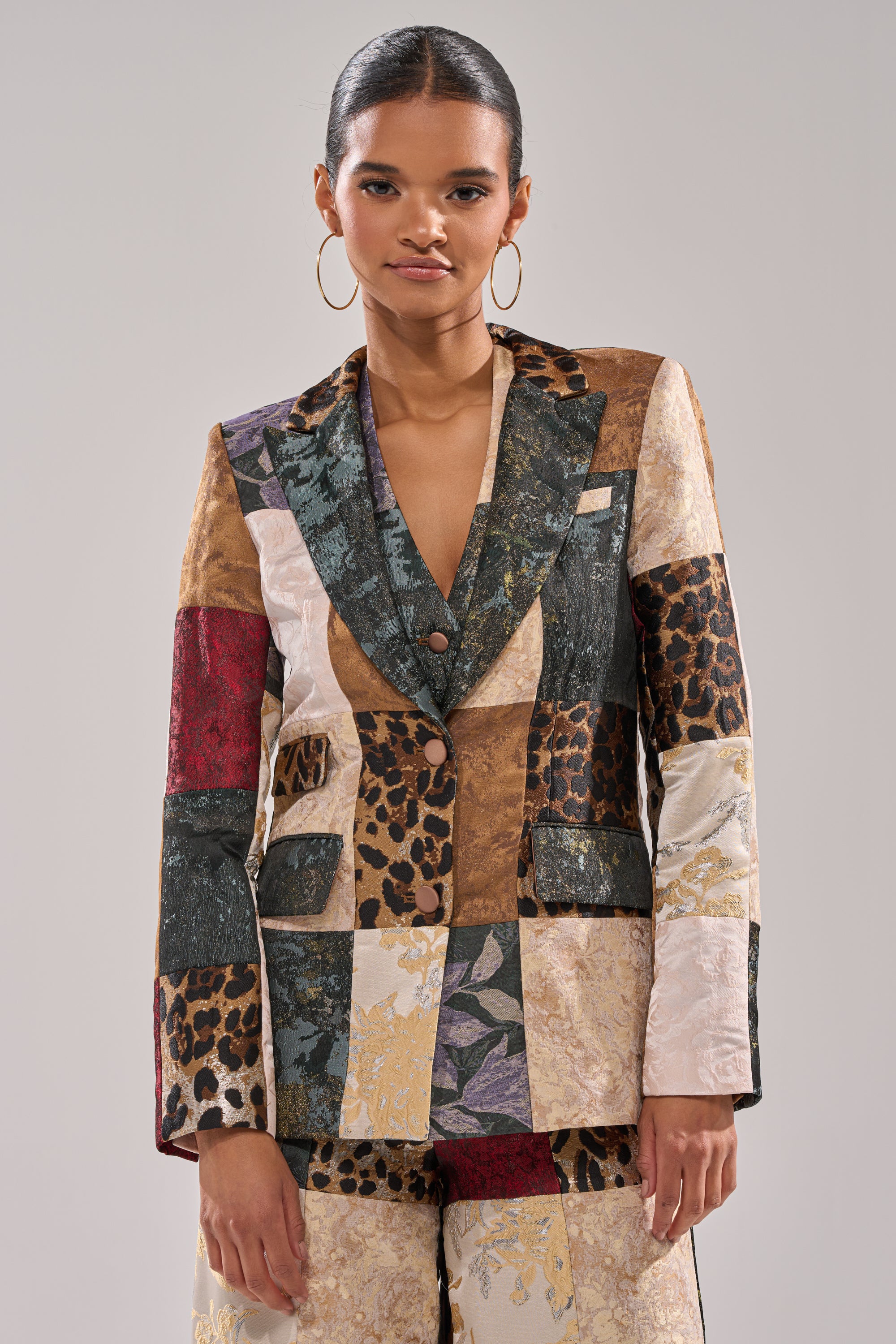 A woman with slicked-back hair models the MADE YOU LOOK PATCHWORK BLAZER, featuring floral, metallic, and animal prints. She stands confidently against a neutral background, accessorized with large hoop earrings.