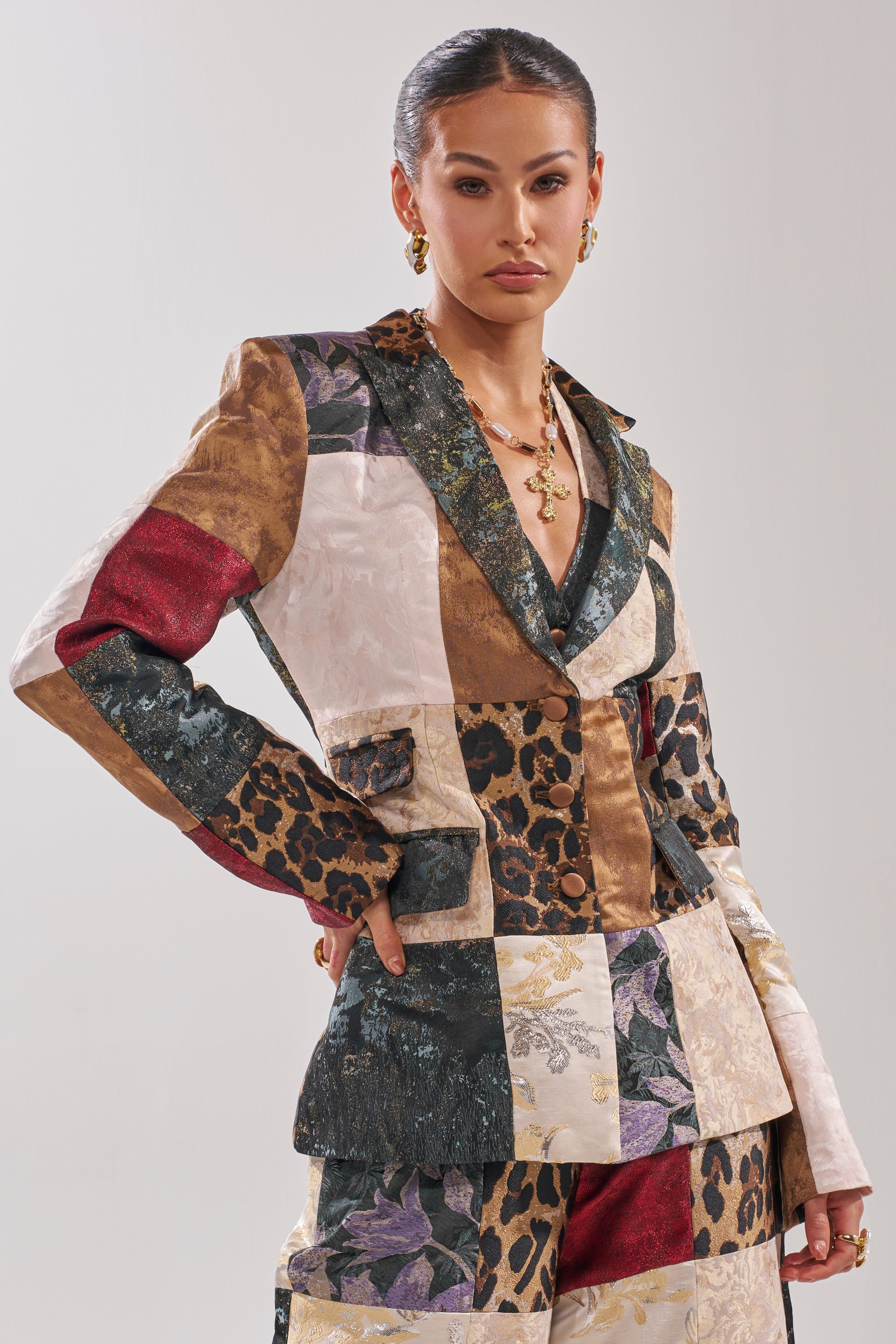 A woman poses confidently in the MADE YOU LOOK PATCHWORK BLAZER, a tailored piece with mixed-media patterns like leopard print, florals, and metallics. She accessorizes with gold jewelry and wears her hair slicked back against a neutral background.