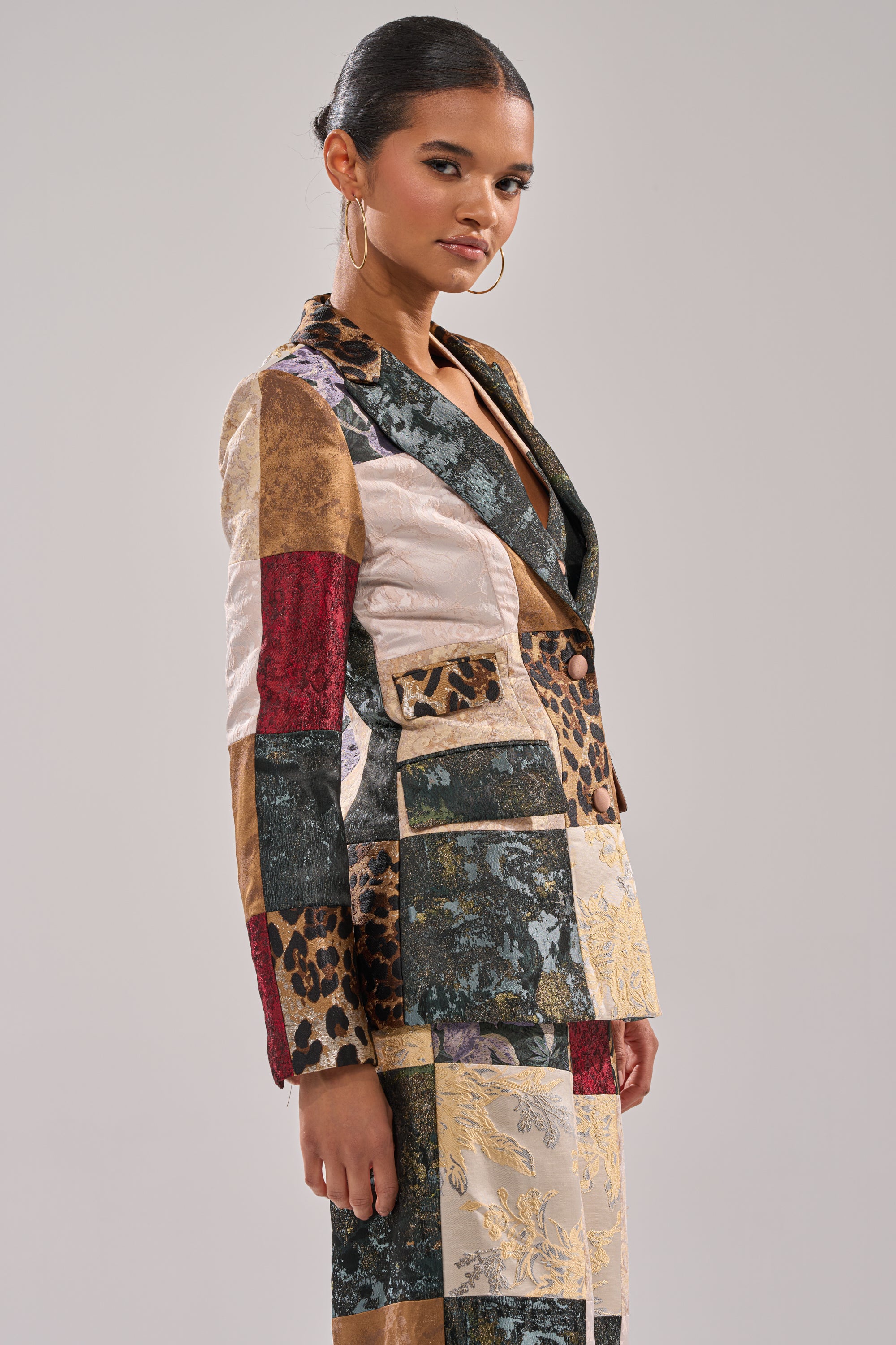 A woman with slicked-back hair and hoop earrings models the MADE YOU LOOK PATCHWORK BLAZER, featuring floral and animal prints, against a plain light gray background.