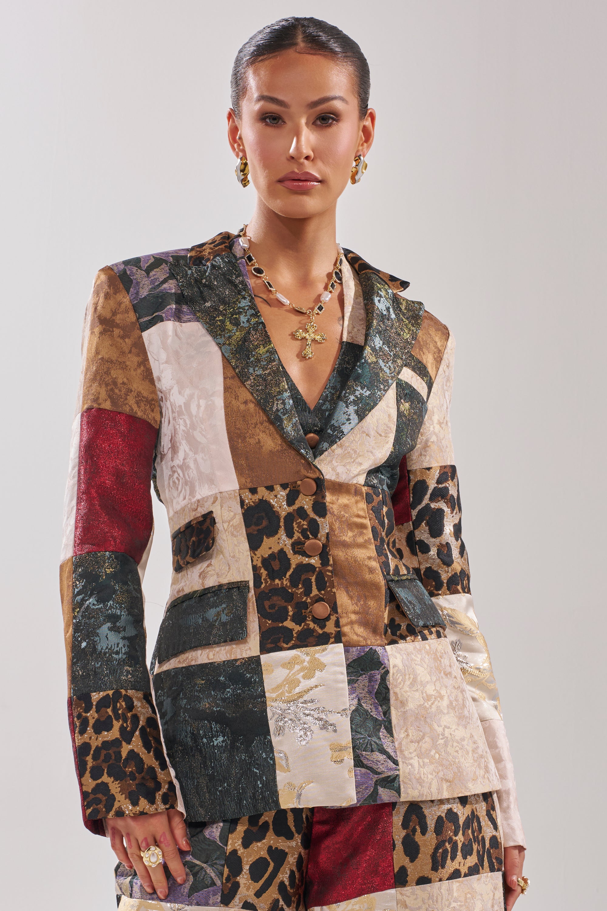 A woman poses against a plain background wearing the MADE YOU LOOK PATCHWORK BLAZER, featuring floral, animal prints, and textured fabrics. She completes her sophisticated look with slicked-back hair and gold jewelry.