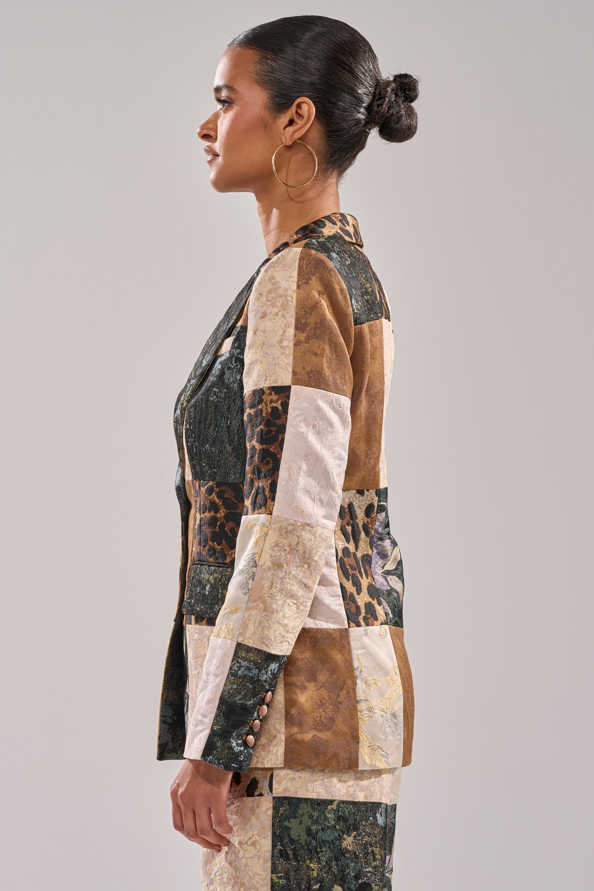 A woman with her hair in a bun and large hoop earrings models the MADE YOU LOOK PATCHWORK BLAZER, showcasing earth tones and animal prints, as she stands in profile against a plain background.
