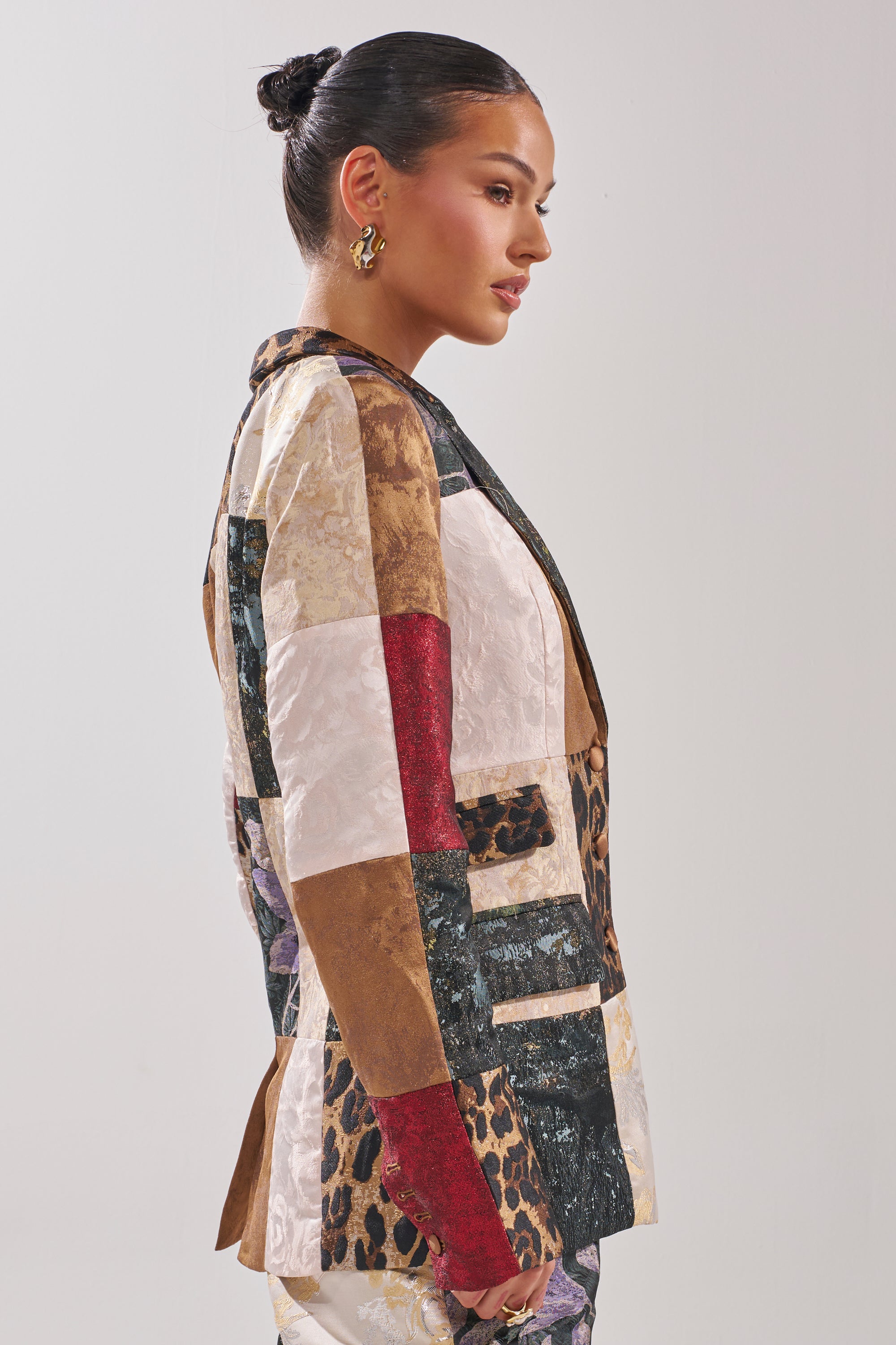 A woman with dark hair in a bun wears gold hoops and the MADE YOU LOOK PATCHWORK BLAZER, featuring leopard print plus red, gold, cream, and black squares, while standing in profile against a plain background.