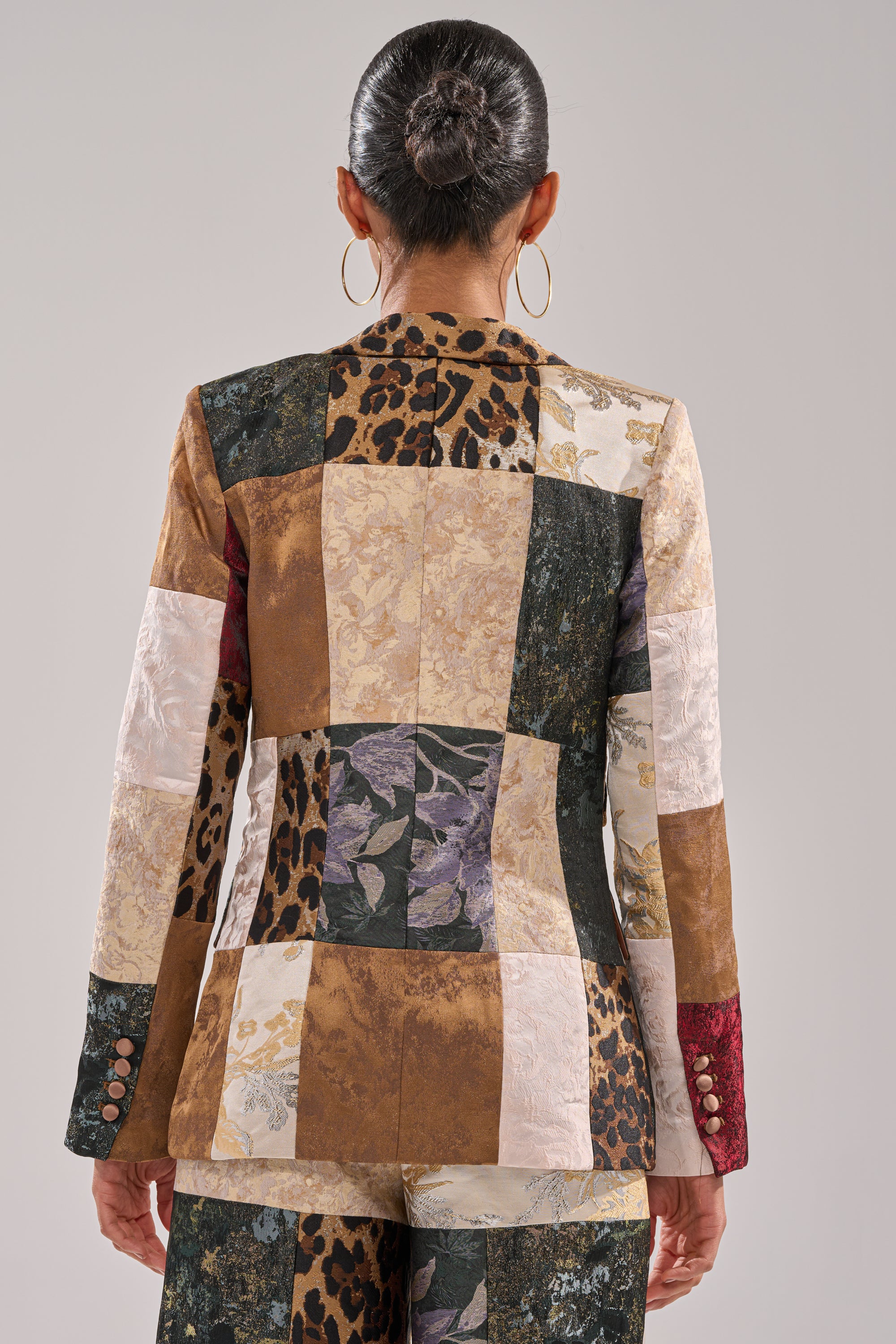 A person with dark hair in a bun and large hoop earrings wears the MADE YOU LOOK PATCHWORK BLAZER, a mixed-media tailored jacket and pants of patterned fabric squares, seen from behind against a plain background.