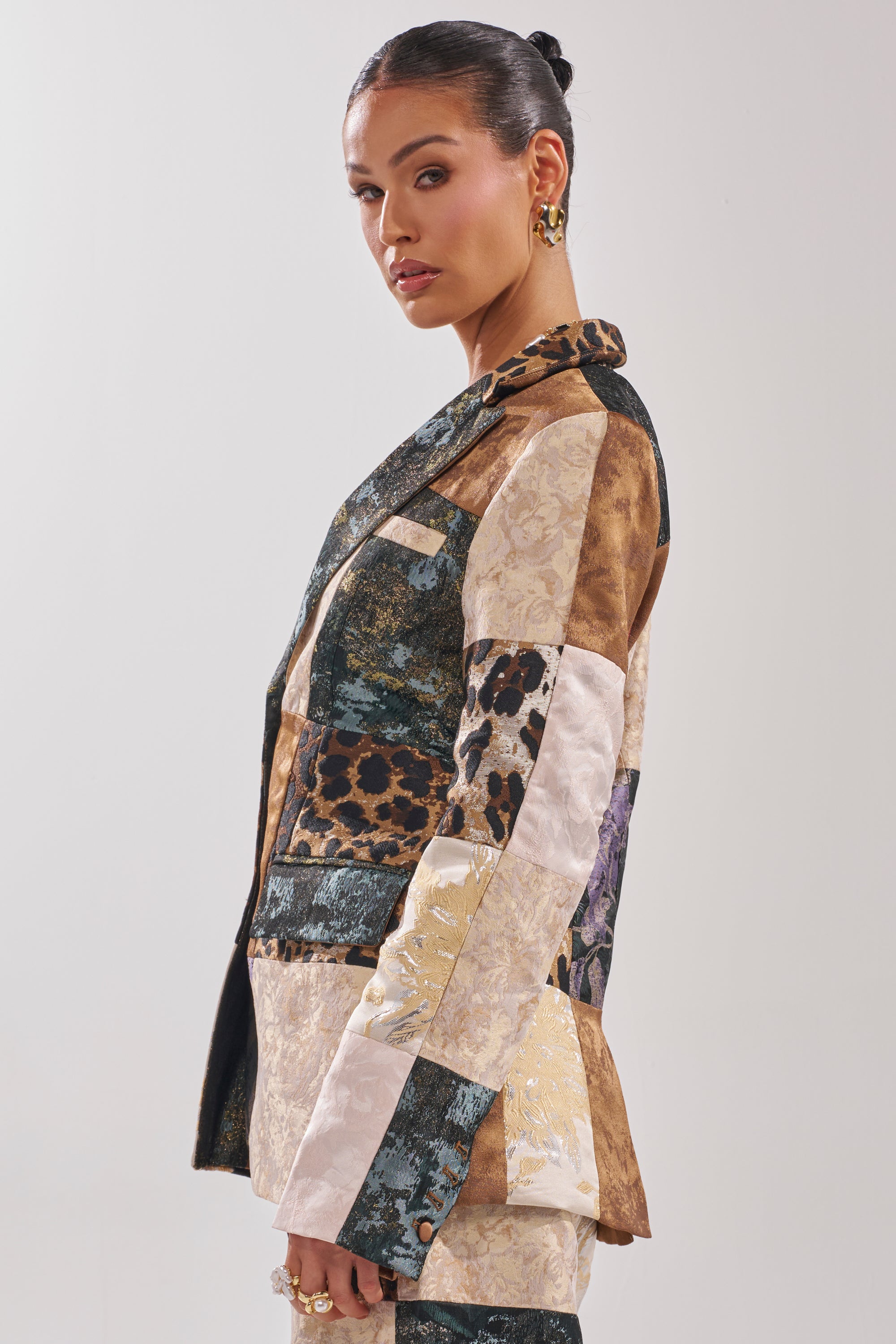 A woman in the MADE YOU LOOK PATCHWORK BLAZER, featuring mixed-media textures, animal prints, and florals, stands in profile against a plain light background with slicked-back hair and gold hoop earrings.