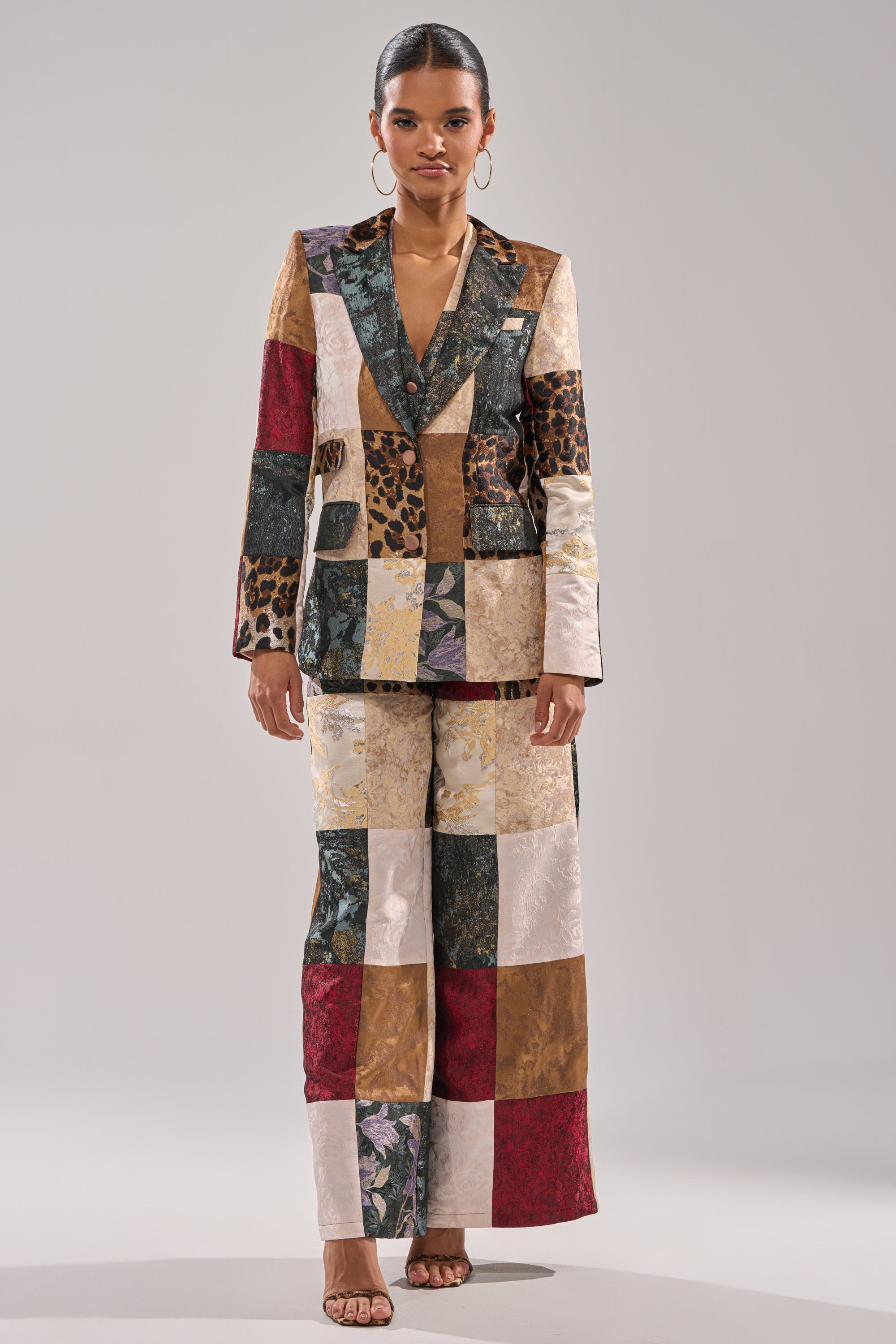 A woman faces forward, wearing the MADE YOU LOOK PATCHWORK BLAZER—a tailored jacket crafted from mixed-media fabrics in colorful floral and animal prints. She completes the look with hoop earrings, neutral heels, and has her hair pulled back.