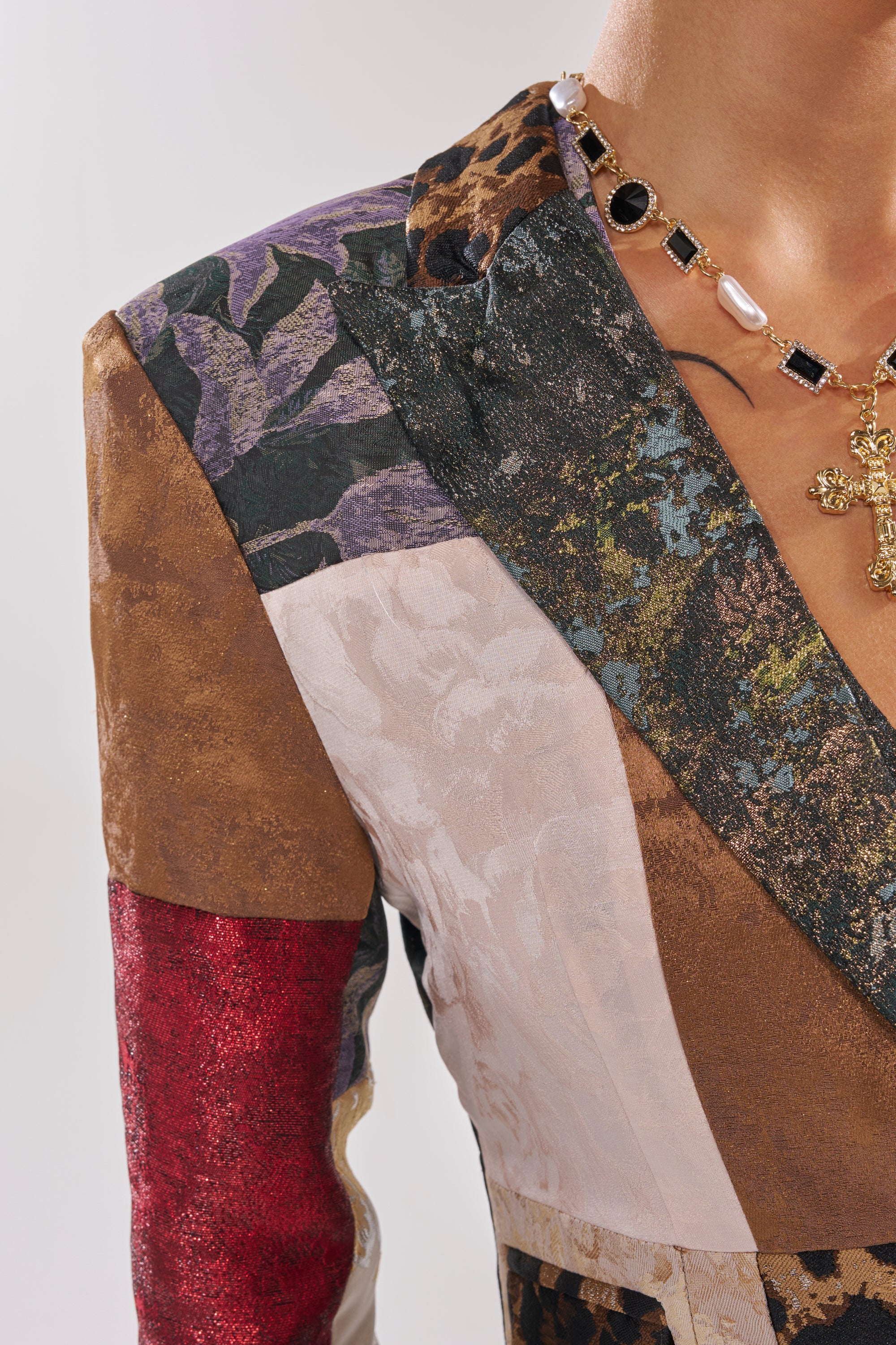 A close-up of a person’s upper body in the MADE YOU LOOK PATCHWORK BLAZER, featuring mixed textures and colors, styled with a gold chain necklace adorned with pearls, black stones, and a cross pendant.