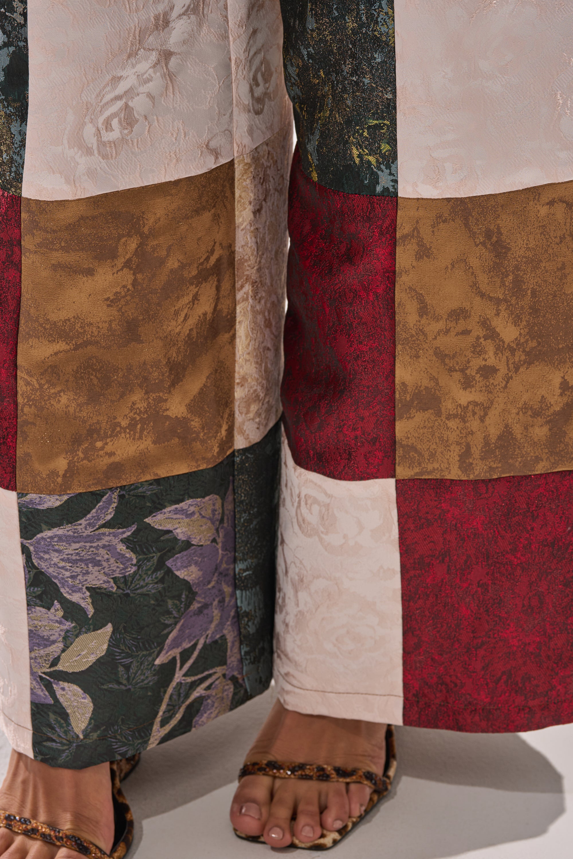 Close-up of a person's feet in sandals on a white surface, wearing the MADE YOU LOOK PATCHWORK TROUSER—high-rise pants featuring red, brown, white, and floral patterned fabric squares.