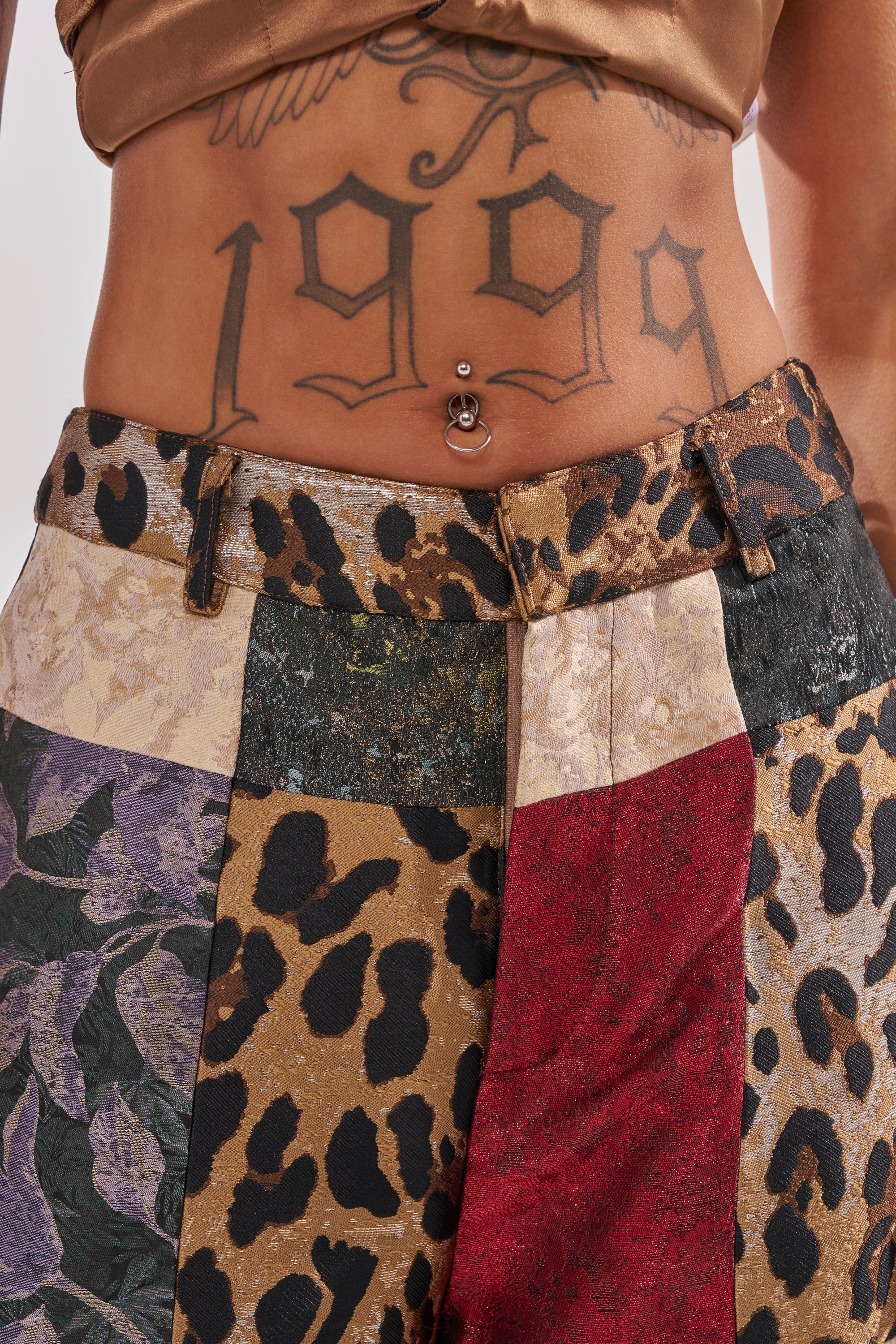 A close-up of a person's midsection showcasing a "1999" tattoo, belly button piercing, and the MADE YOU LOOK PATCHWORK TROUSER with a mix of animal prints and unique fabrics.