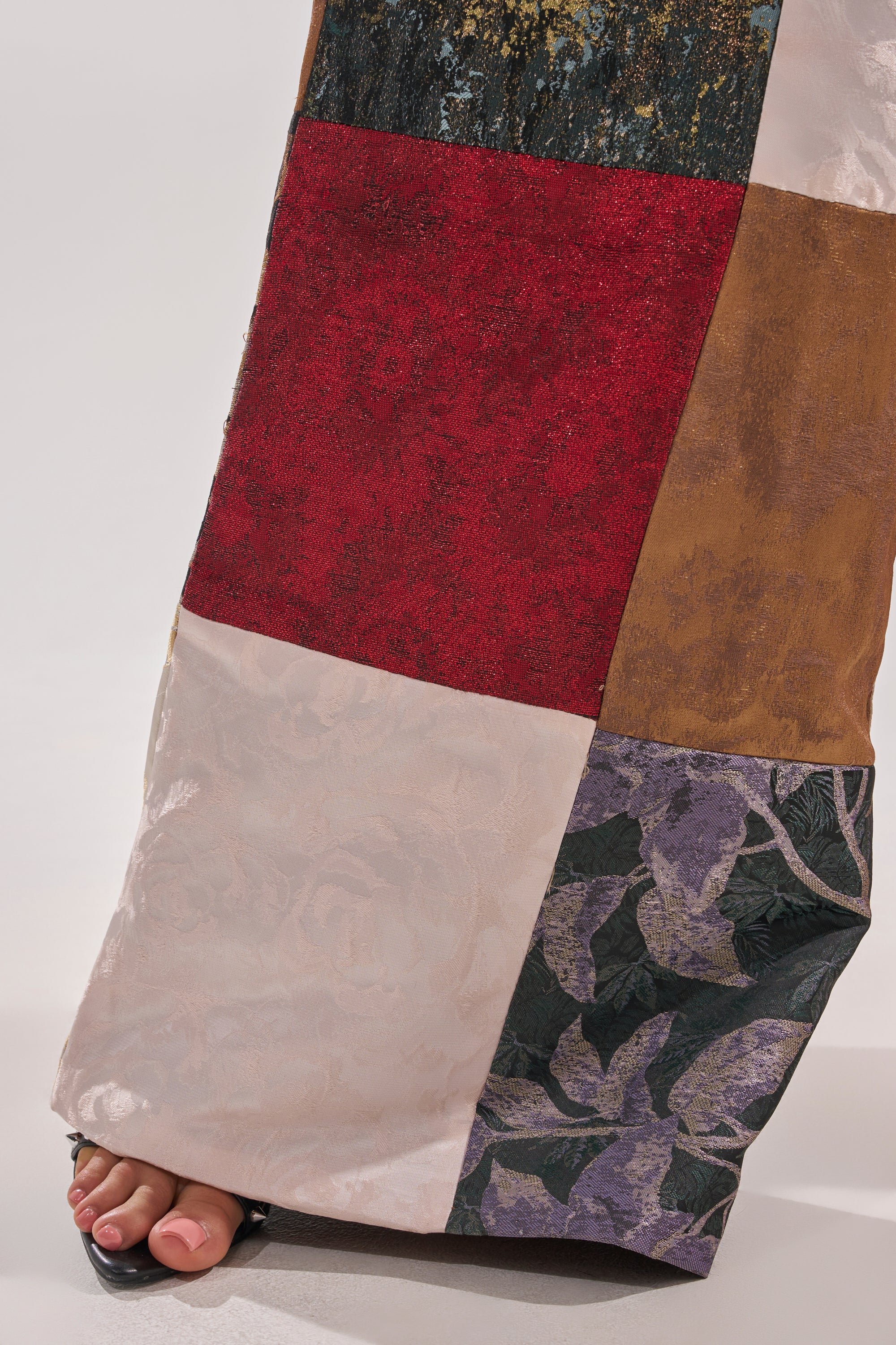 A close-up of the MADE YOU LOOK PATCHWORK TROUSER features colorful fabric squares in various textures, with a glimpse of bare toes and part of a sandal at the bottom left.