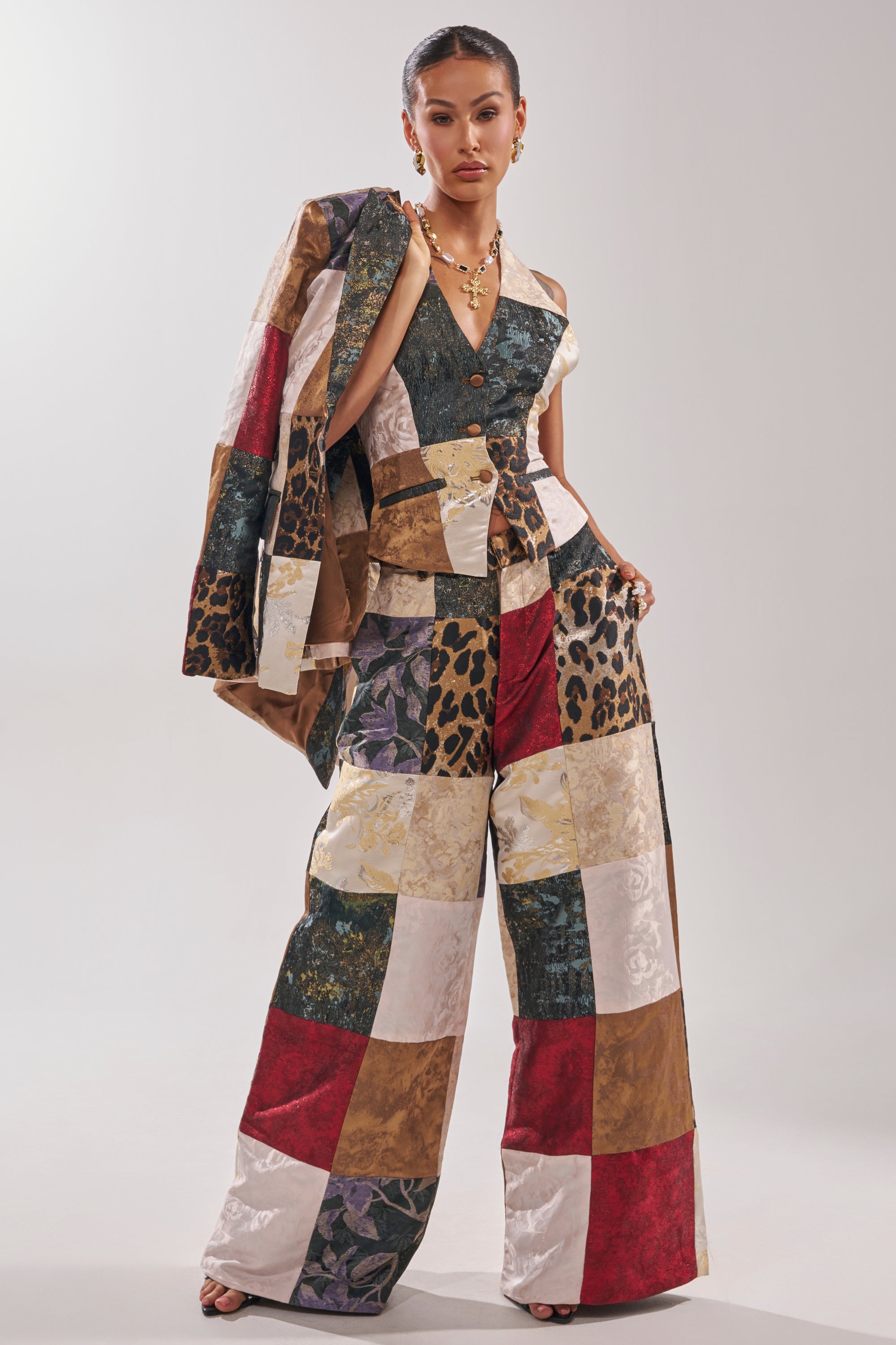 A woman poses confidently in the MADE YOU LOOK PATCHWORK TROUSER, featuring wide-leg, high-rise pants with red, white, gold, green, and animal print squares. She completes the look with statement jewelry and a slicked-back hairstyle.