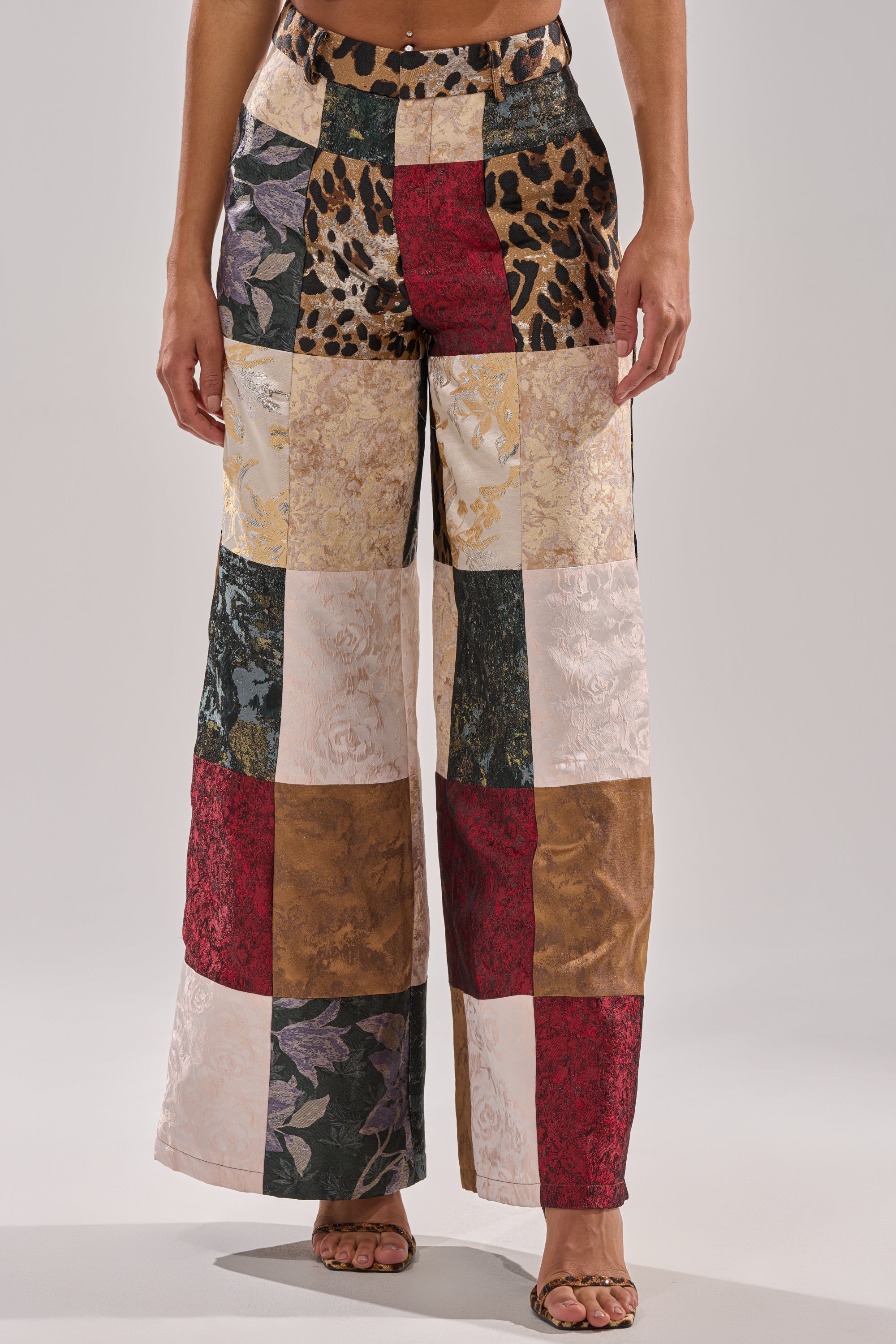 A person wearing the MADE YOU LOOK PATCHWORK TROUSER—high-rise wide-leg pants with colorful brocade jacquard, floral, animal print, and solid fabric squares—paired with open-toe sandals.