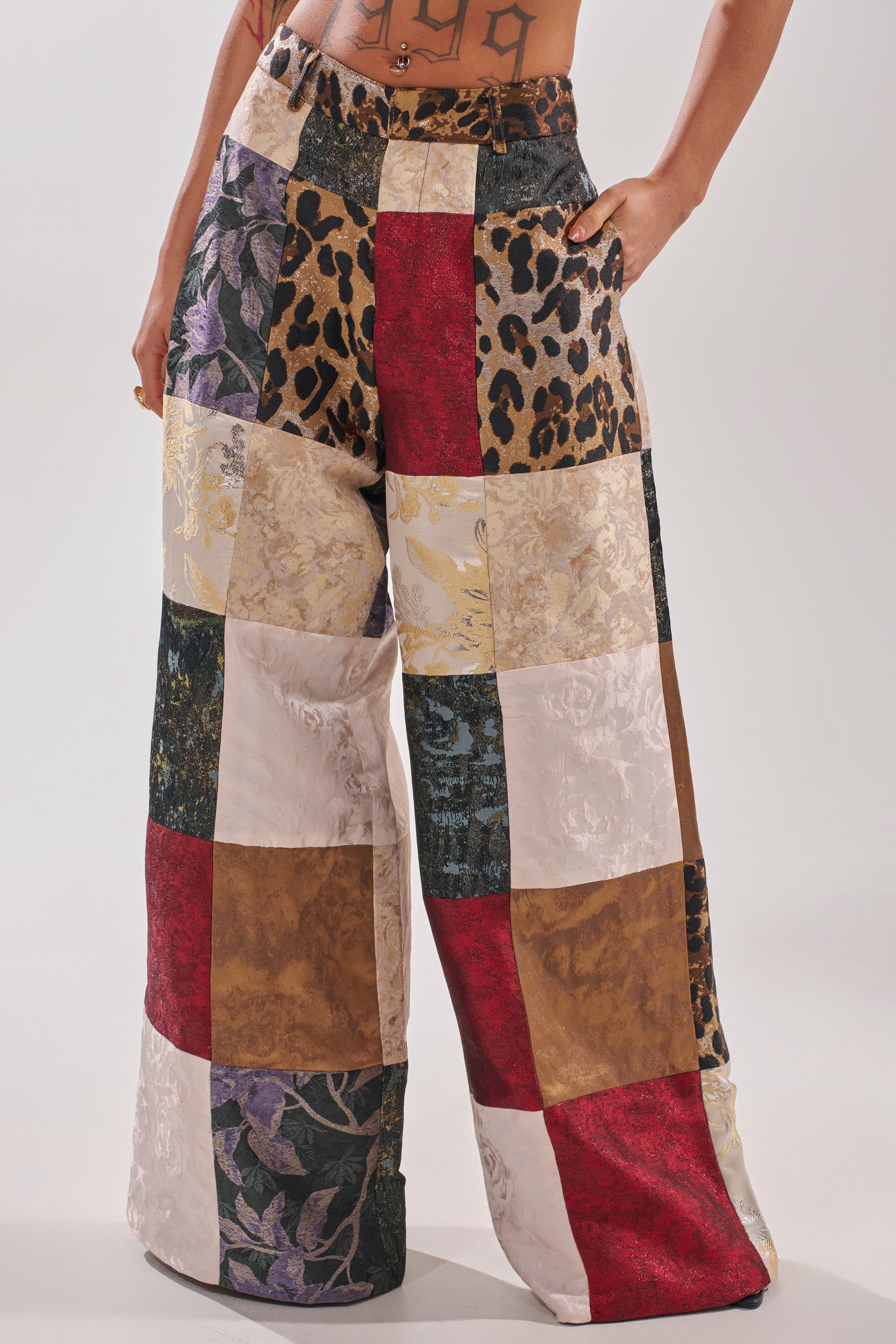 Person wearing MADE YOU LOOK PATCHWORK TROUSER with wide-leg fit and textured fabric squares in florals, animal print, metallic gold, red, cream, and black. Only the lower torso and legs are visible.