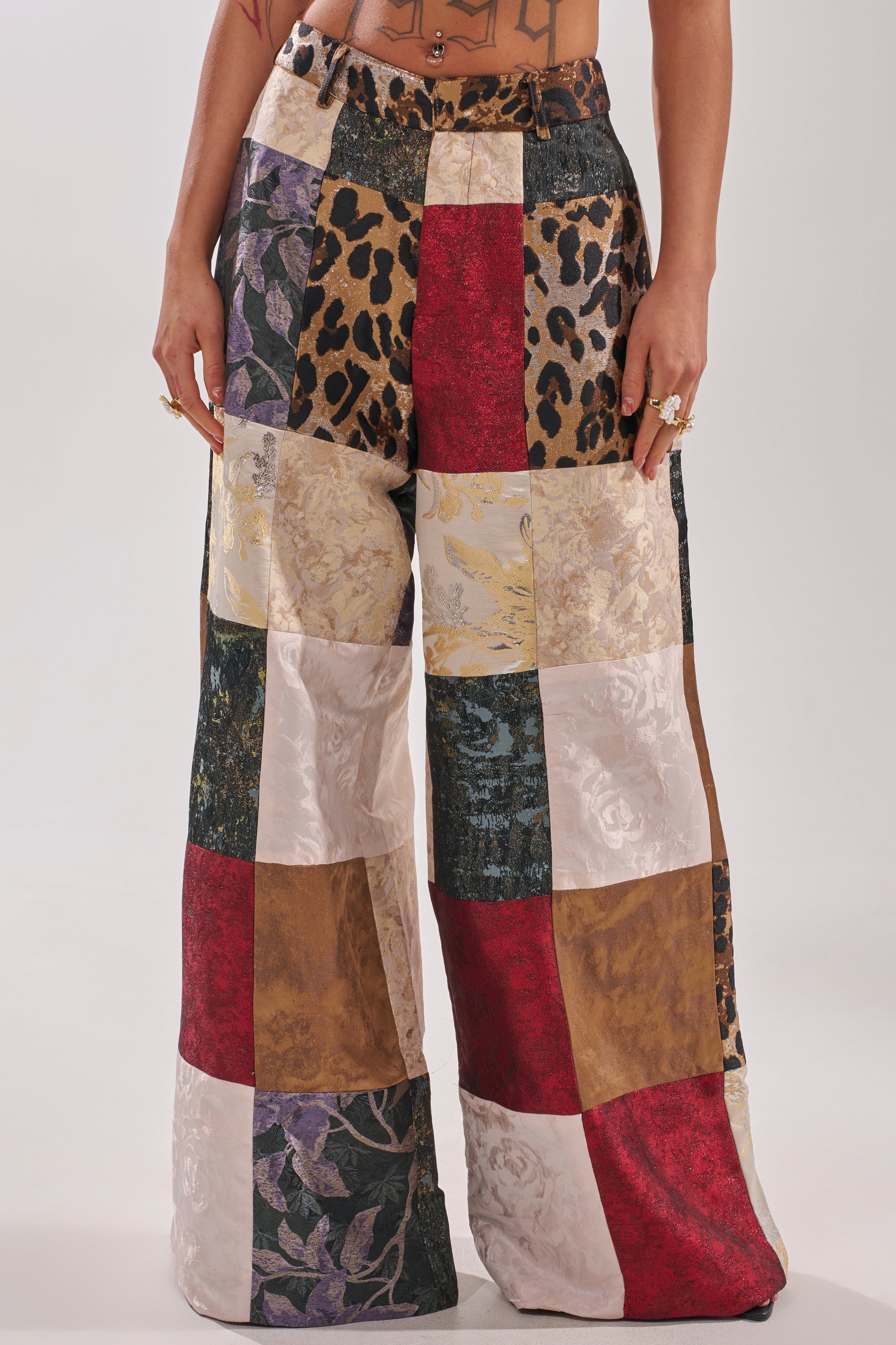 Wearing the MADE YOU LOOK PATCHWORK TROUSER, a person showcases high-rise, wide-leg pants made from textured squares in animal print, floral, gold, red, black, and white—highlighting the unique silhouette from lower torso to legs.