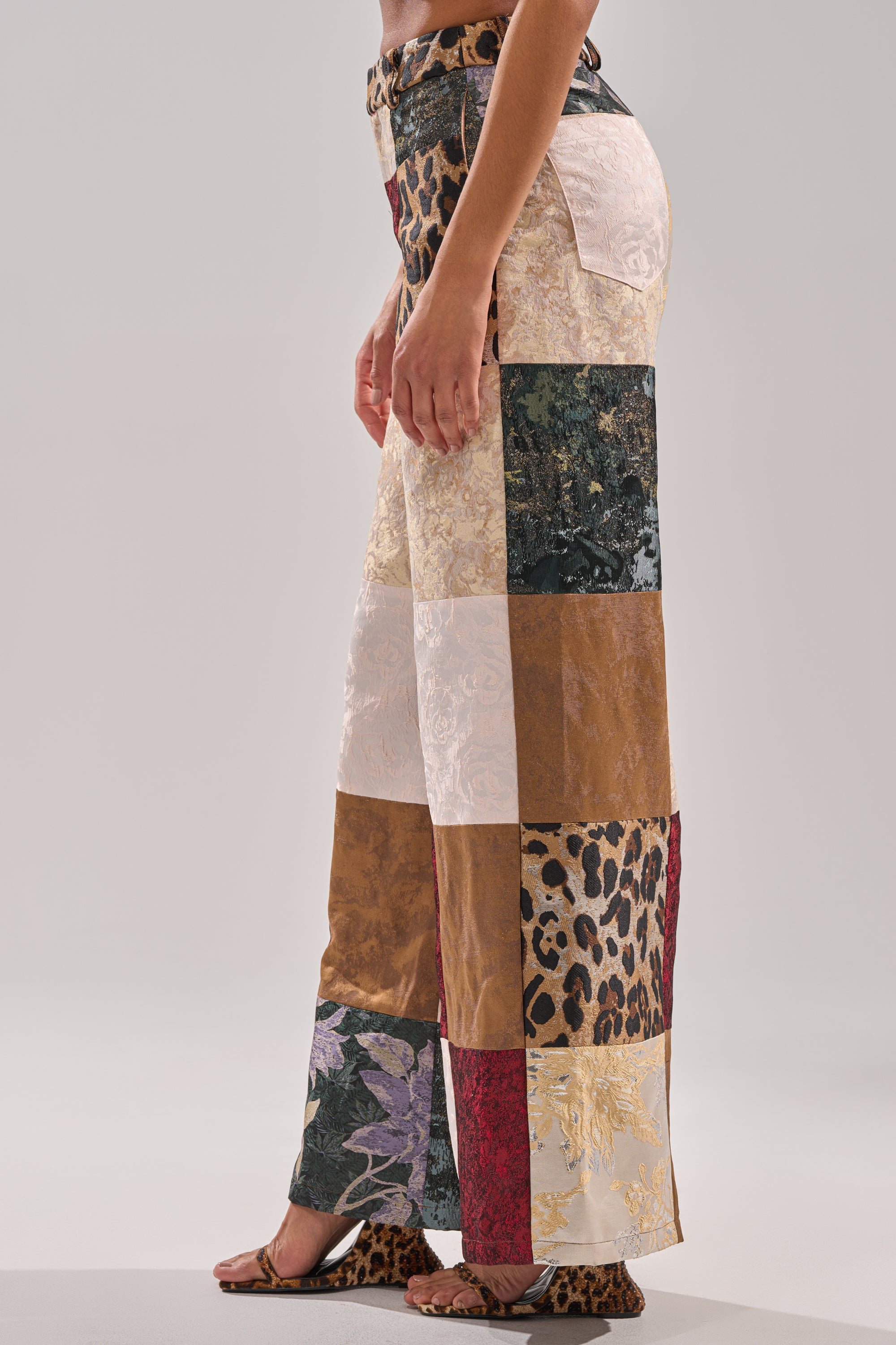 A person stands sideways wearing the MADE YOU LOOK PATCHWORK TROUSER—high-rise, wide-leg pants with mixed patterns and textures like leopard print, florals, suede, and brocade—paired with open-toe sandals that also have a leopard print.