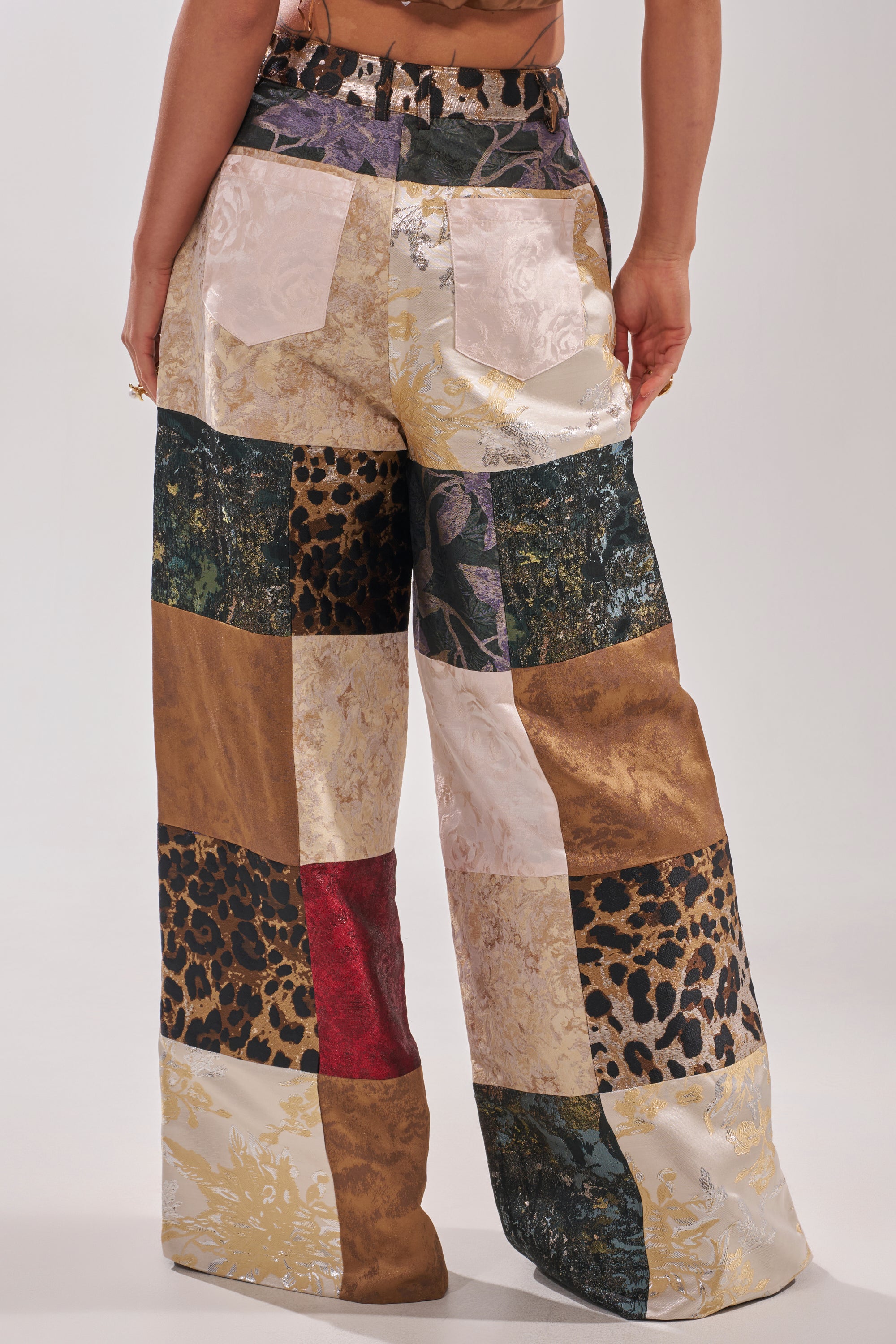 A person faces away, wearing the MADE YOU LOOK PATCHWORK TROUSER—high-rise, wide-leg pants crafted from textured and patterned fabrics with animal prints, florals, metallics, and earth-toned muted colors.