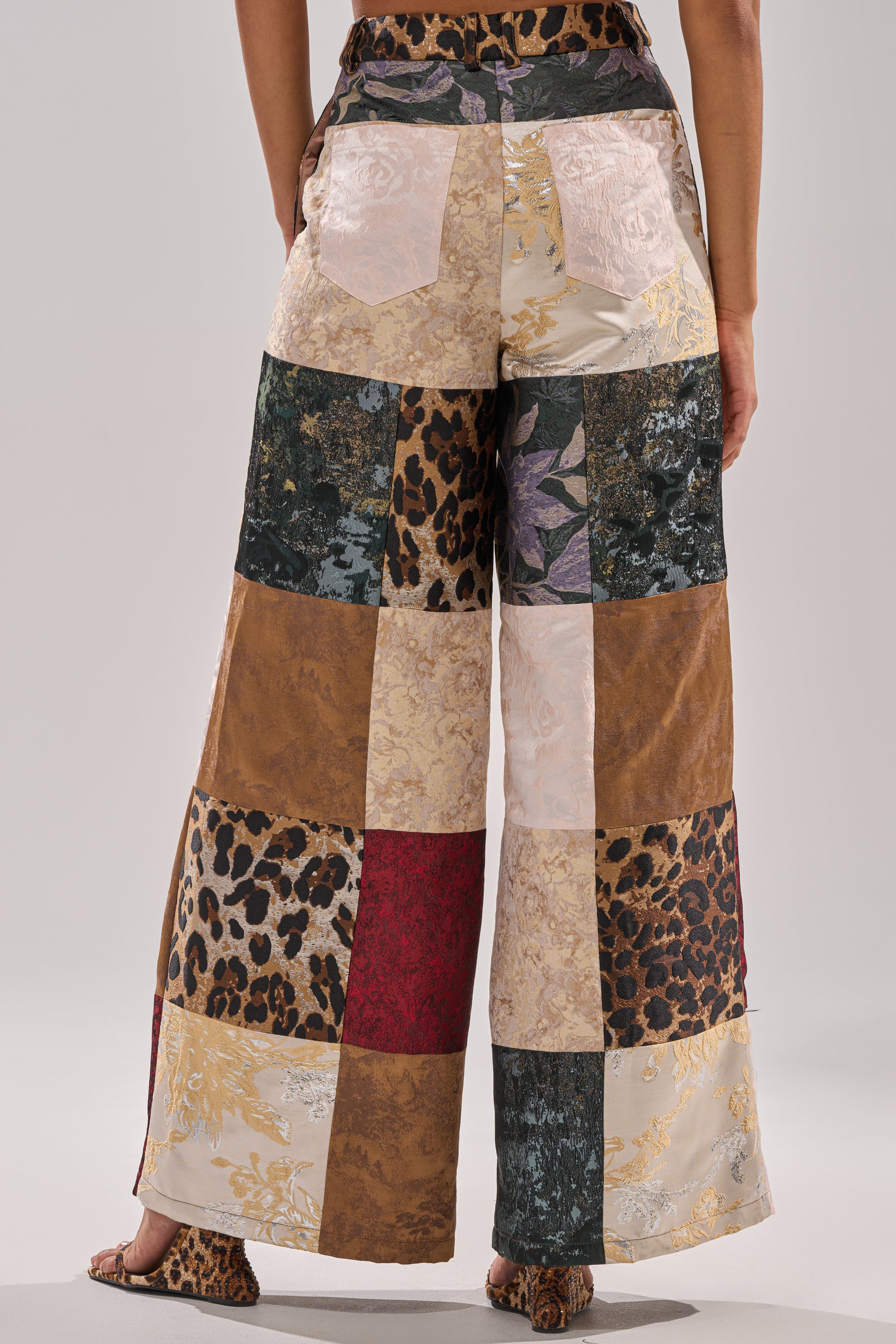 Someone stands with their back to the camera wearing the MADE YOU LOOK PATCHWORK TROUSER—wide-leg, high-rise pants crafted from various patterned and textured fabric squares, including animal prints and florals.