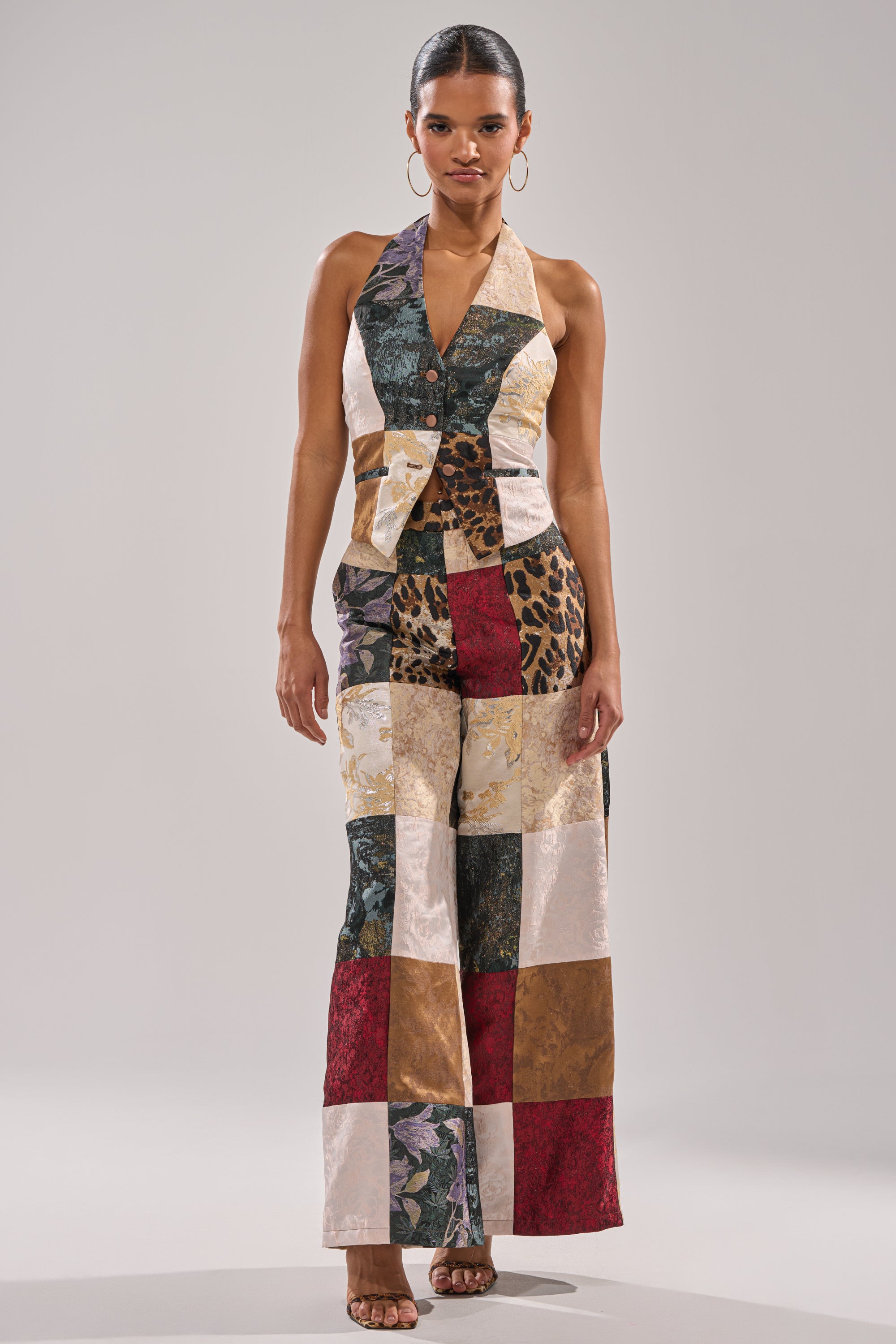 A woman models the MADE YOU LOOK PATCHWORK TROUSER, a sleeveless halter jumpsuit featuring wide legs and a patchwork of floral, leopard print, and solid color squares. She accessorizes with hoop earrings and open-toe heels.