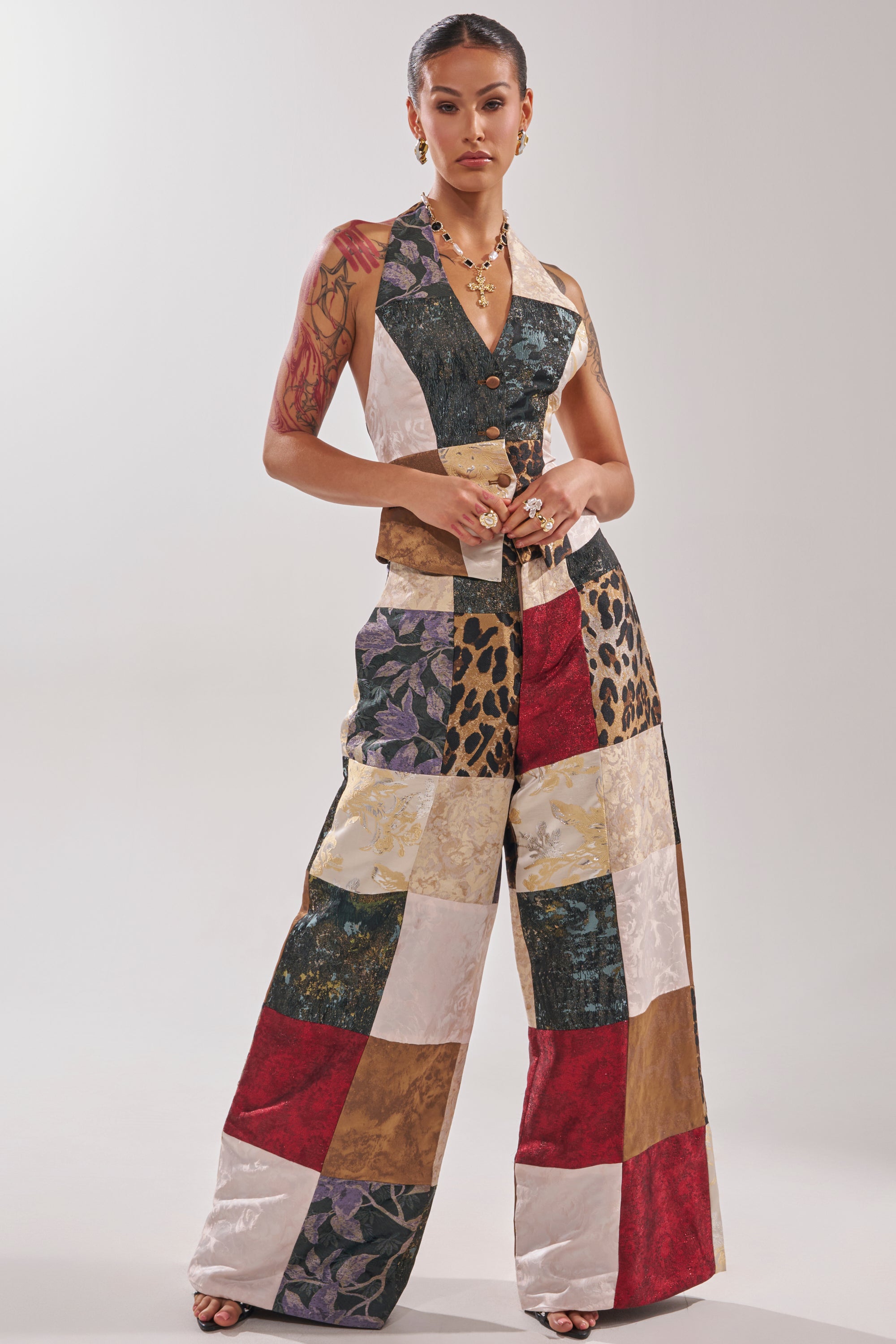 A woman exudes confidence in the MADE YOU LOOK PATCHWORK TROUSER, a high-rise, wide-leg pant with colorful floral, leopard, and solid prints. She pairs it with a matching vest, accessorizes with jewelry, and shows off an upper arm tattoo.