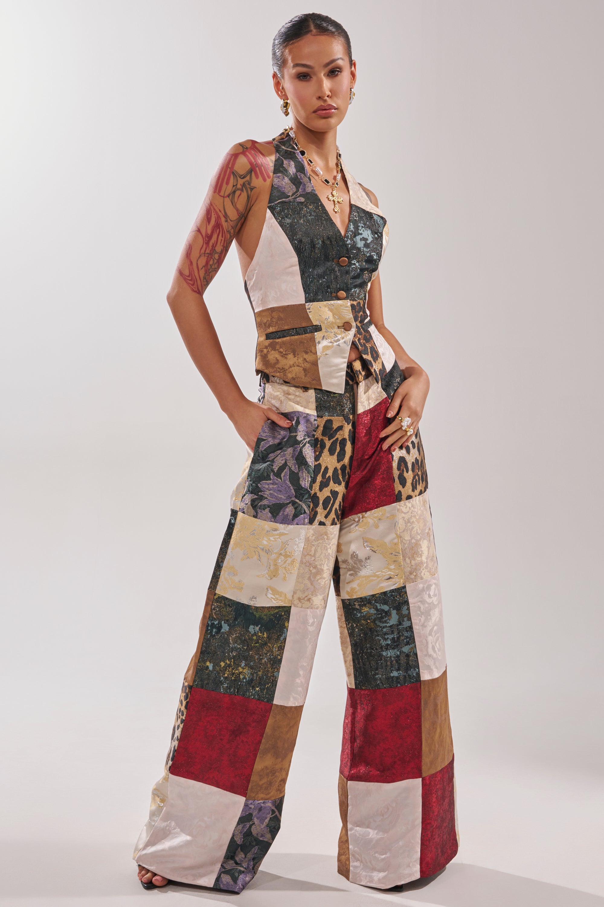 A woman stands confidently in the MADE YOU LOOK PATCHWORK TROUSER, featuring wide legs and a bold patchwork of animal prints, florals, and vibrant colors. She shows off visible tattoos and accessorizes with jewelry.