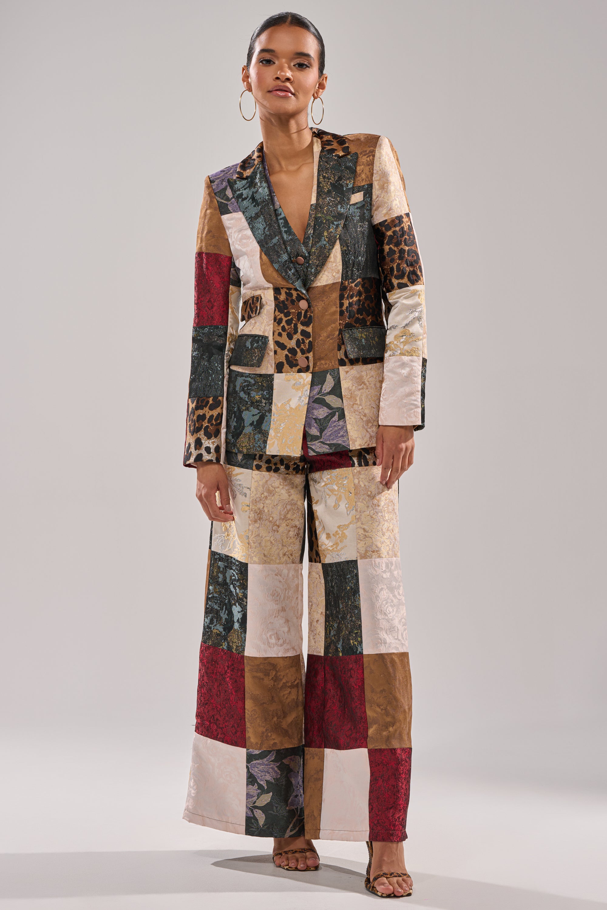 A woman models the MADE YOU LOOK PATCHWORK TROUSER—a wide-leg pant with vibrant floral, animal print, and solid patches—styled with hoop earrings and sandals against a neutral background.