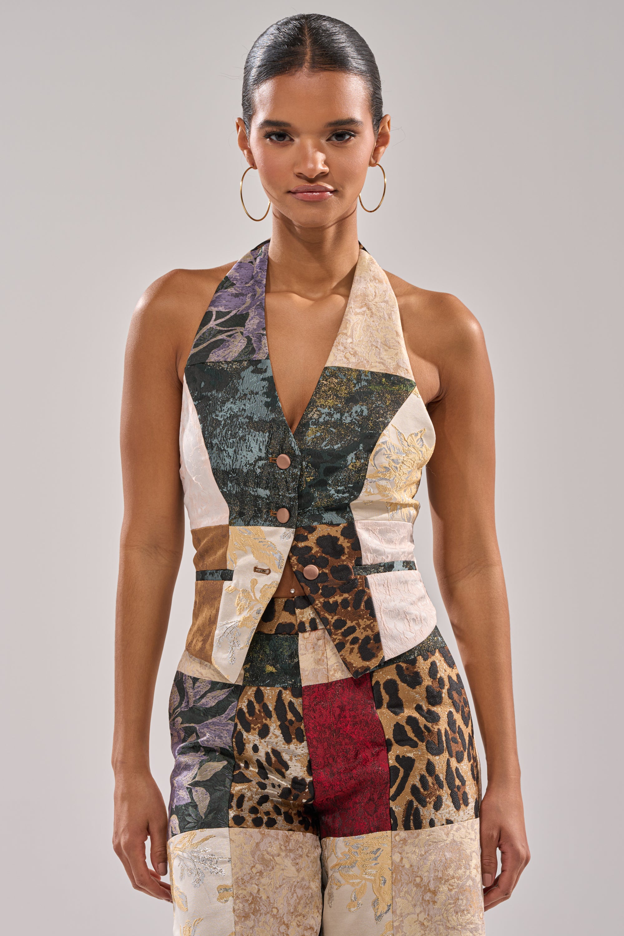 Wearing the MADE YOU LOOK PATCHWORK HALTER VEST and matching pants featuring leopard, floral, and brocade jacquard details, a woman stands against a plain background with hoop earrings and her hair pulled back.