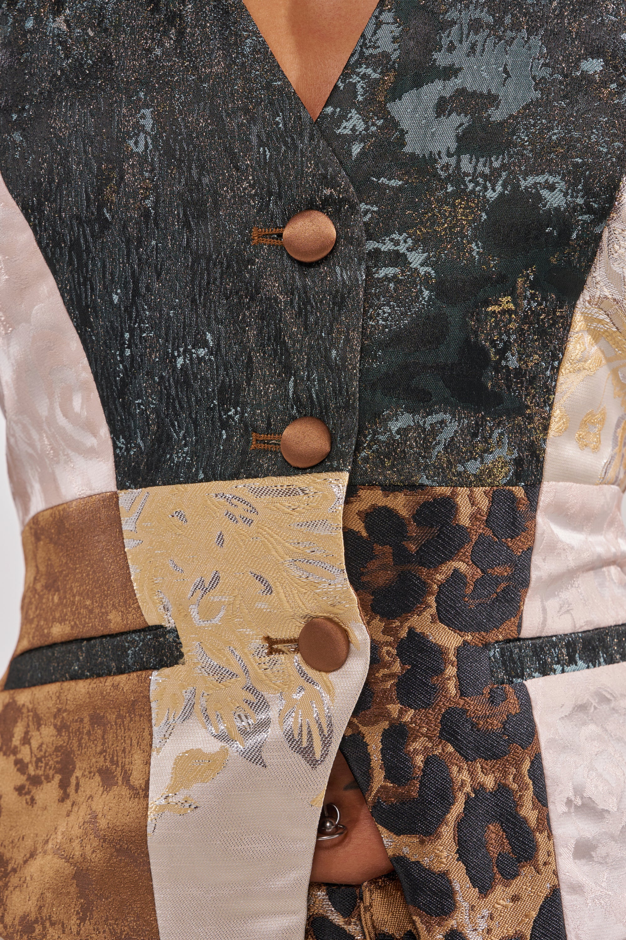 Close-up of a person wearing the MADE YOU LOOK PATCHWORK HALTER VEST featuring textured, patterned fabrics—floral, leopard print, metallic gold, dark green—and fastened with round brown buttons.