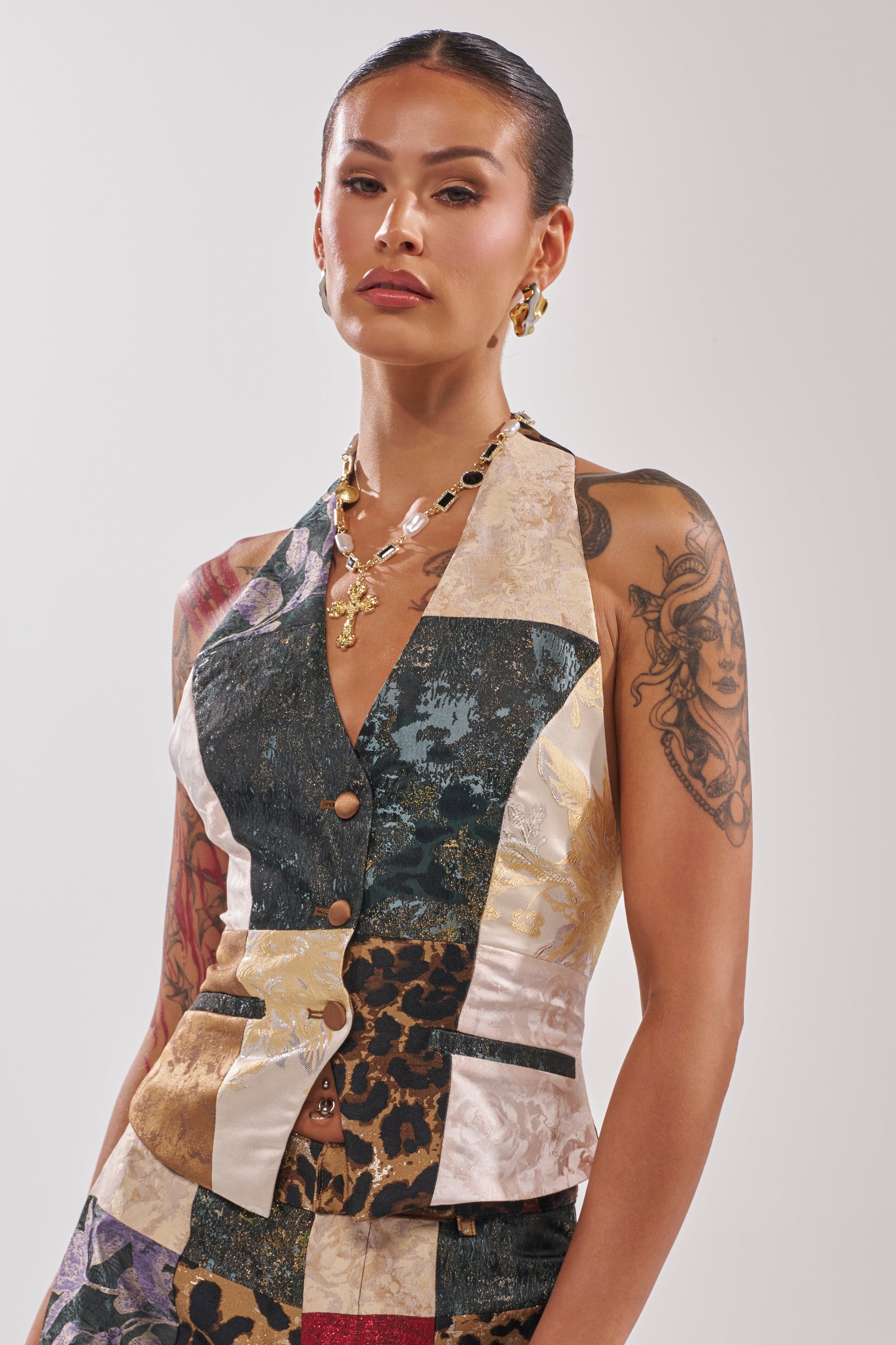A woman with slicked-back hair and visible arm tattoos wears the MADE YOU LOOK PATCHWORK HALTER VEST, styled with gold jewelry, posing against a plain light background.