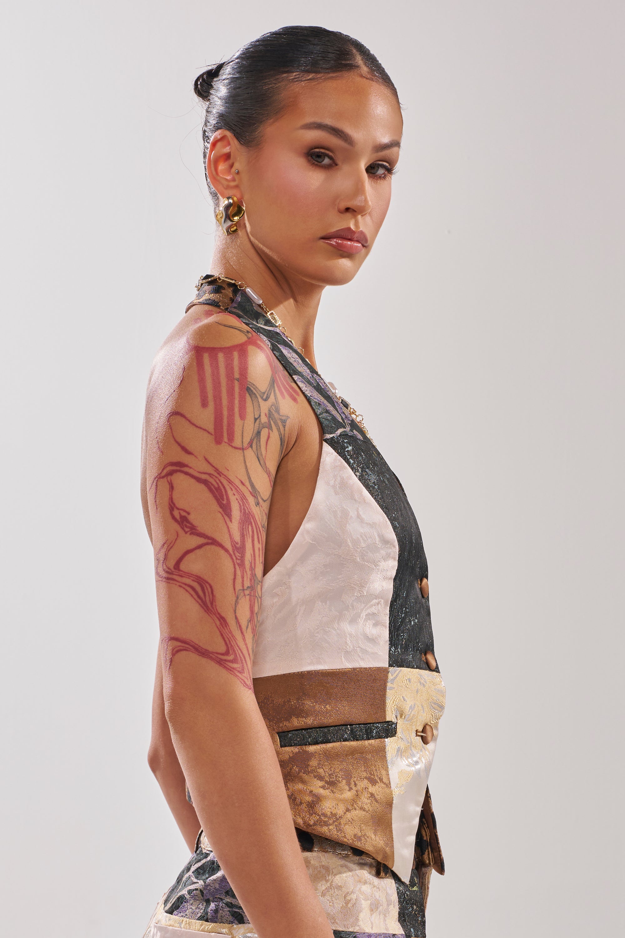 A person with dark hair pulled back, wearing the MADE YOU LOOK PATCHWORK HALTER VEST and gold earrings, stands in profile against a plain background, revealing a prominent red abstract tattoo on their upper arm.