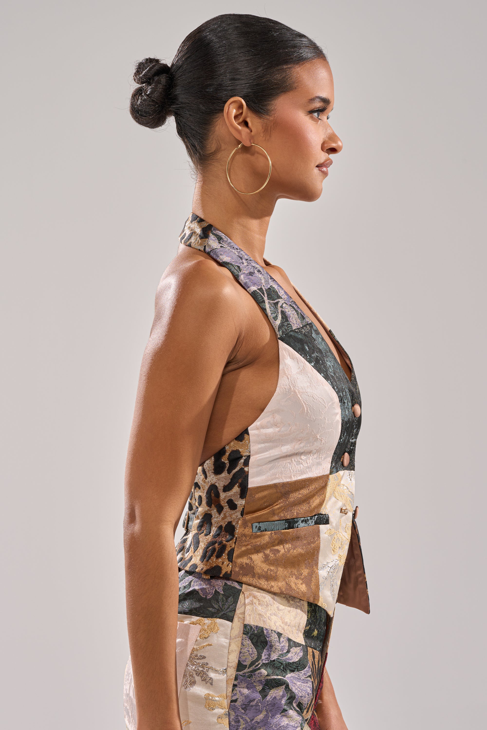 A woman with dark hair in a bun stands in profile, wearing large hoop earrings and the MADE YOU LOOK PATCHWORK HALTER VEST featuring mixed fabrics and prints. The background is plain and light-colored.