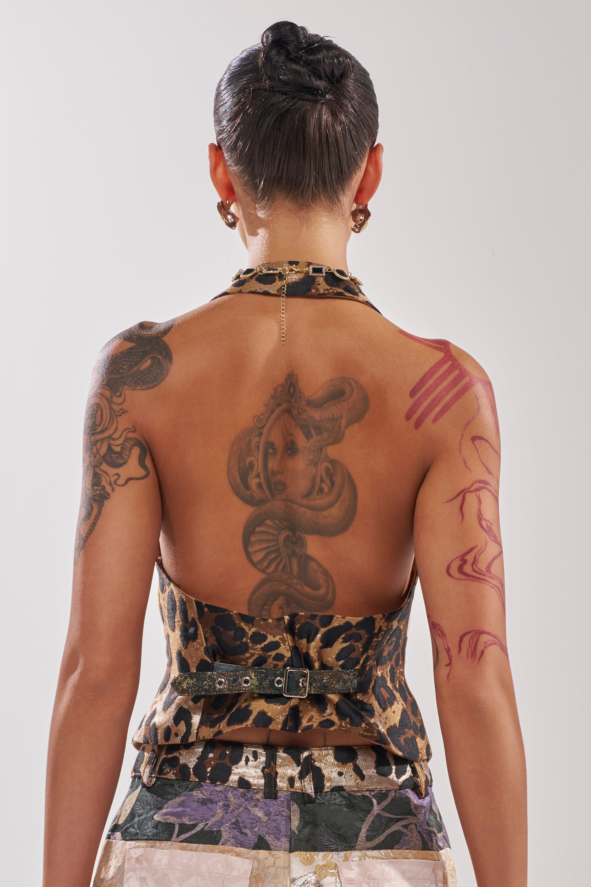 A person with short dark hair is seen from behind wearing the MADE YOU LOOK PATCHWORK HALTER VEST, which features an animal print, halter-style open back. Multiple tattoos, including a large snake on their back, are visible.