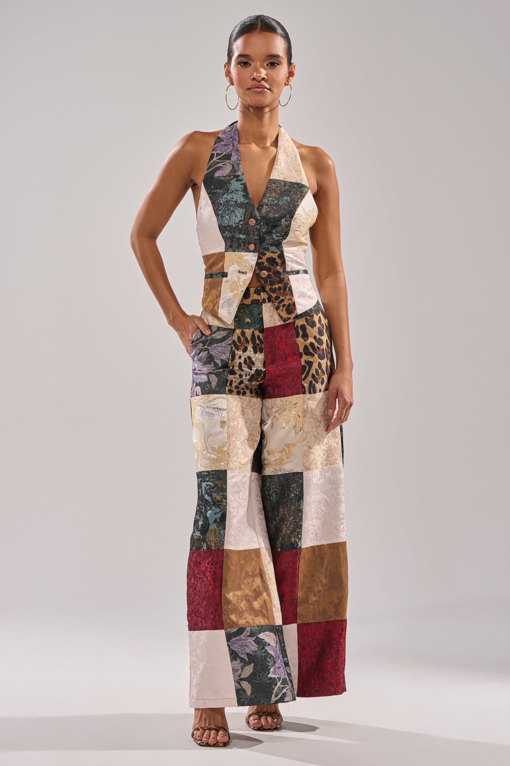 A woman wears the MADE YOU LOOK PATCHWORK HALTER VEST, featuring mixed patterns and colors. She completes her look with hoop earrings and open-toe heels while posing against a neutral background.