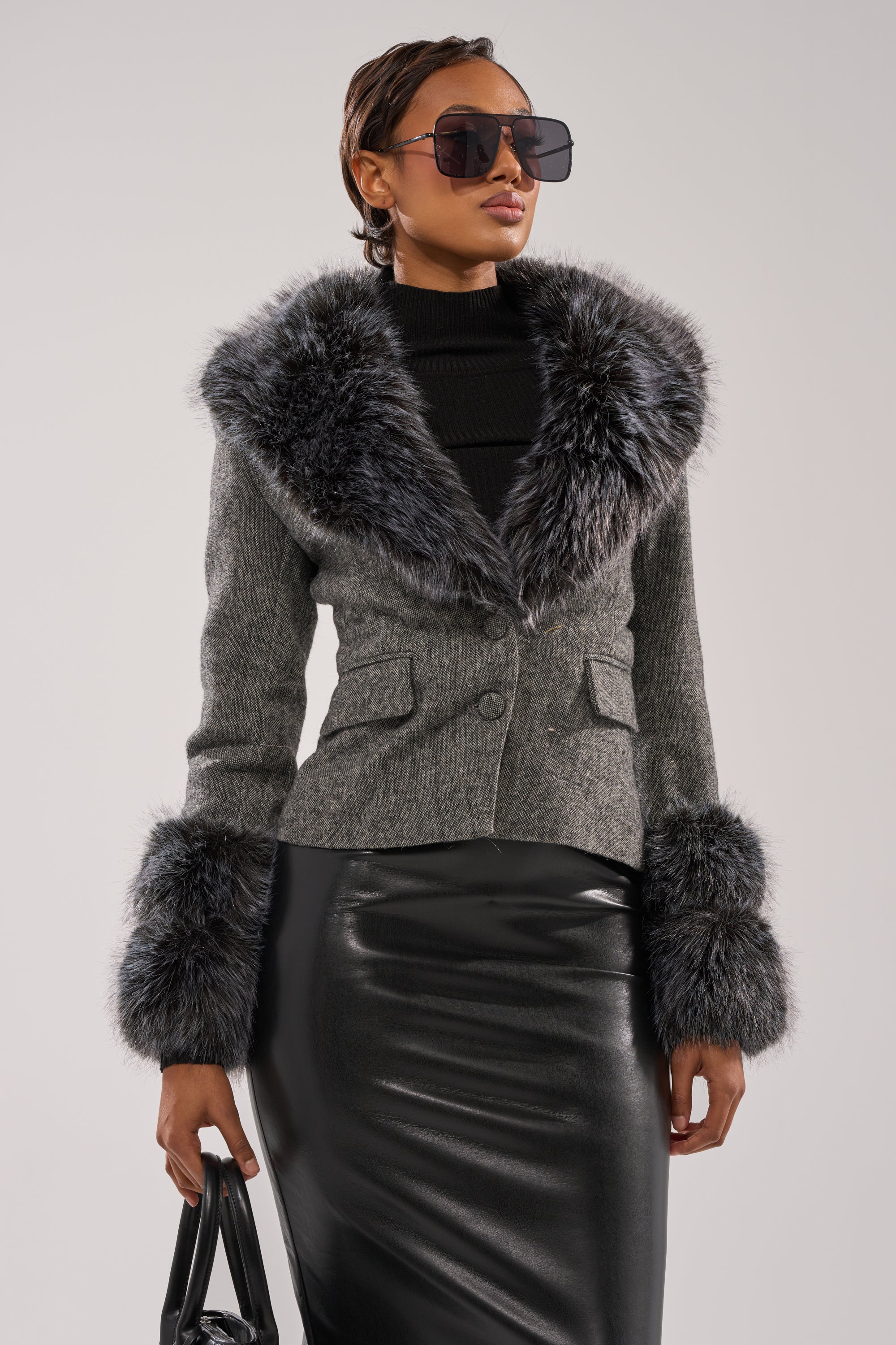 A woman in large sunglasses and the I SEE LONDON FUR TRIM BLAZER, styled with a black turtleneck, shiny black skirt, and black handbag, poses against a neutral background.