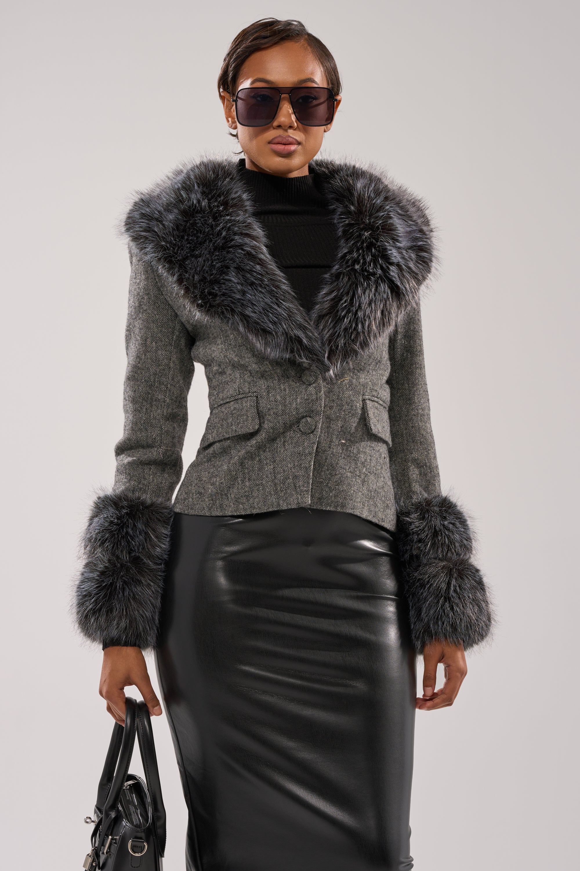 A woman wears large sunglasses, a black turtleneck, a shiny black leather skirt, and the I SEE LONDON FUR TRIM BLAZER with faux fur collar and cuffs, holding a black handbag against a plain background.