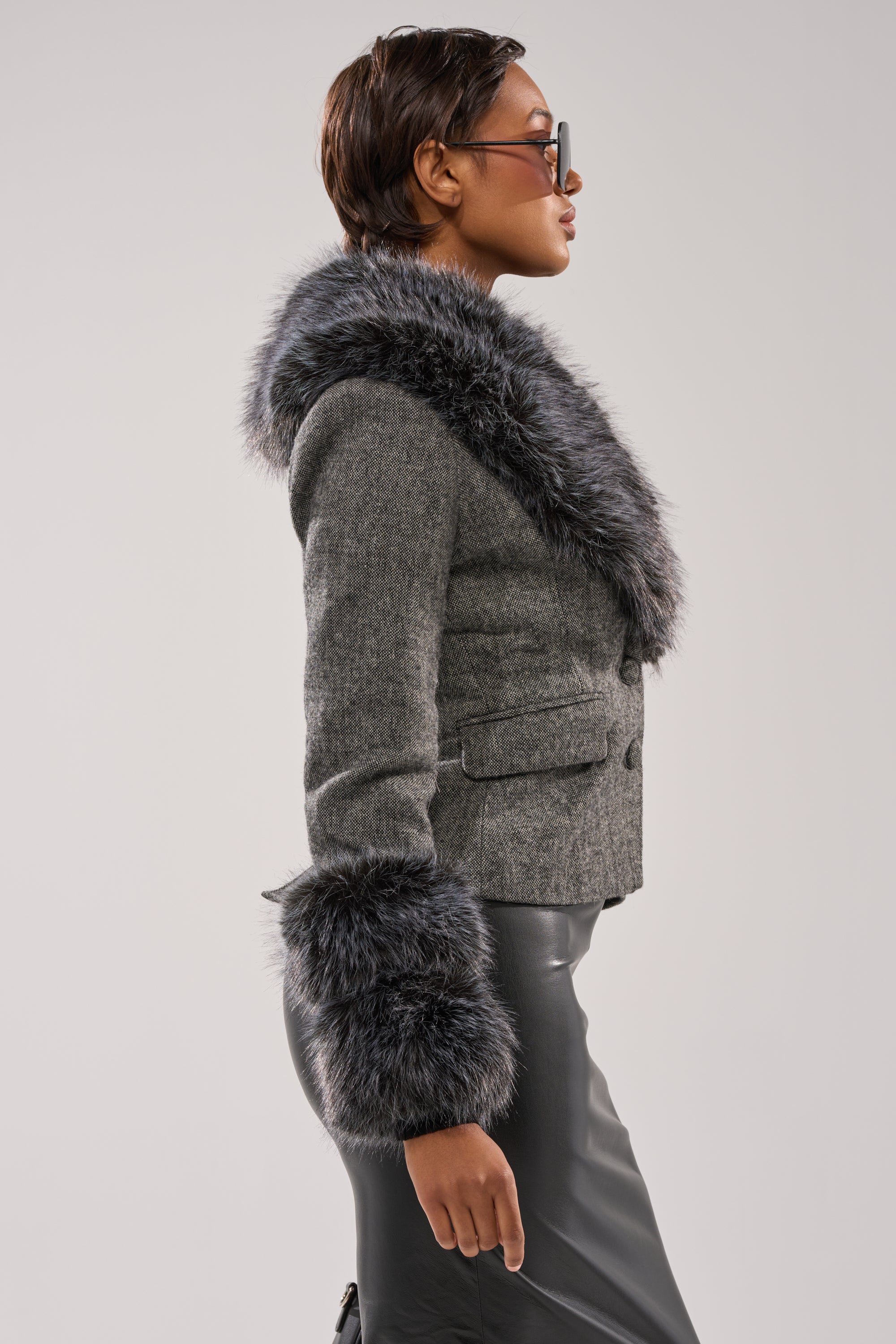 A person in profile wears the I SEE LONDON FUR TRIM BLAZER with a fitted collar and cuffs, paired with sunglasses and a black leather skirt, standing against a plain light background.