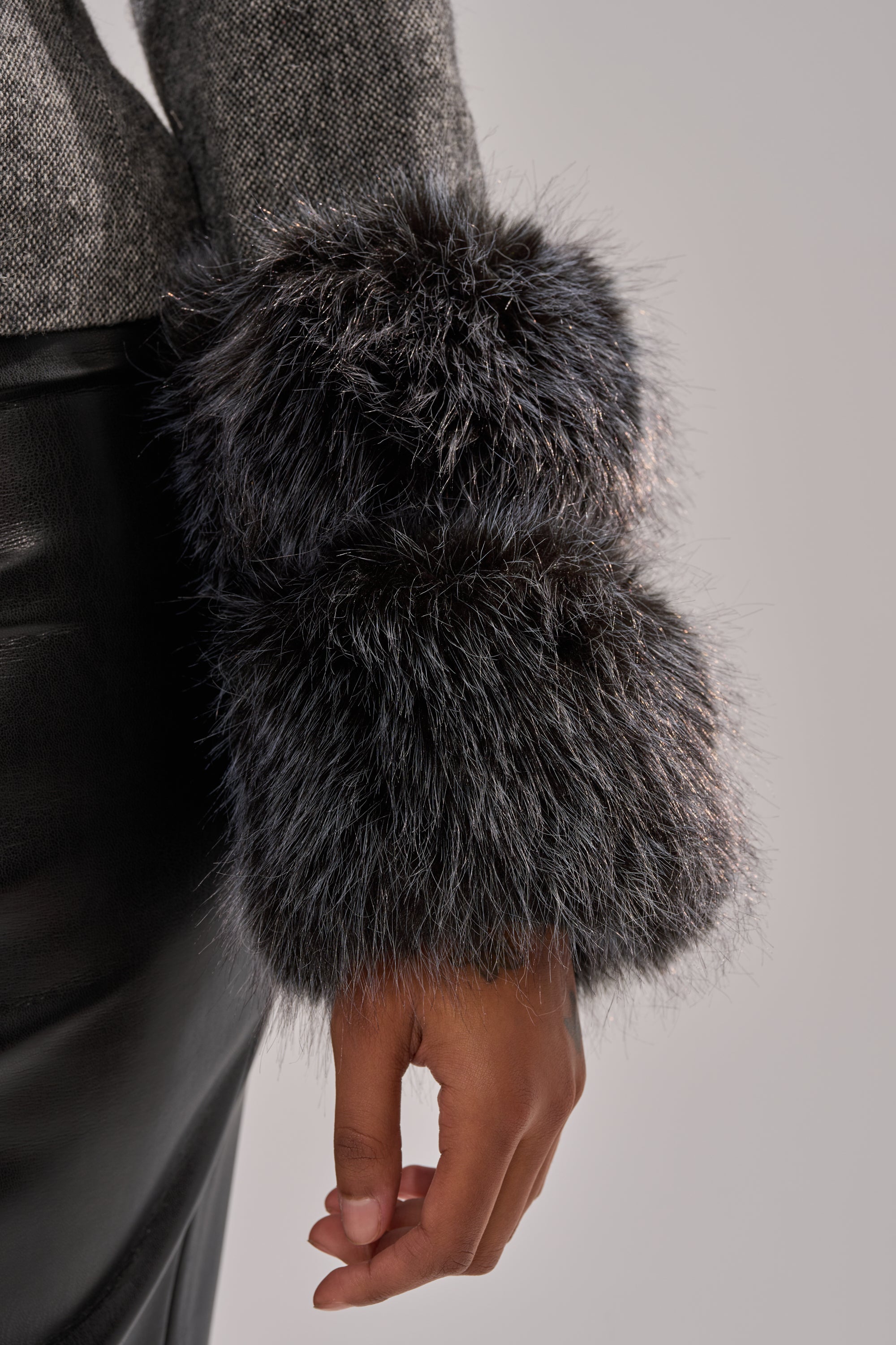 A person wearing the I SEE LONDON FUR TRIM BLAZER, featuring gray wool-blend fabric and thick faux fur cuffs, paired with a black leather skirt; shown from the waist down with their arm relaxed by their side.