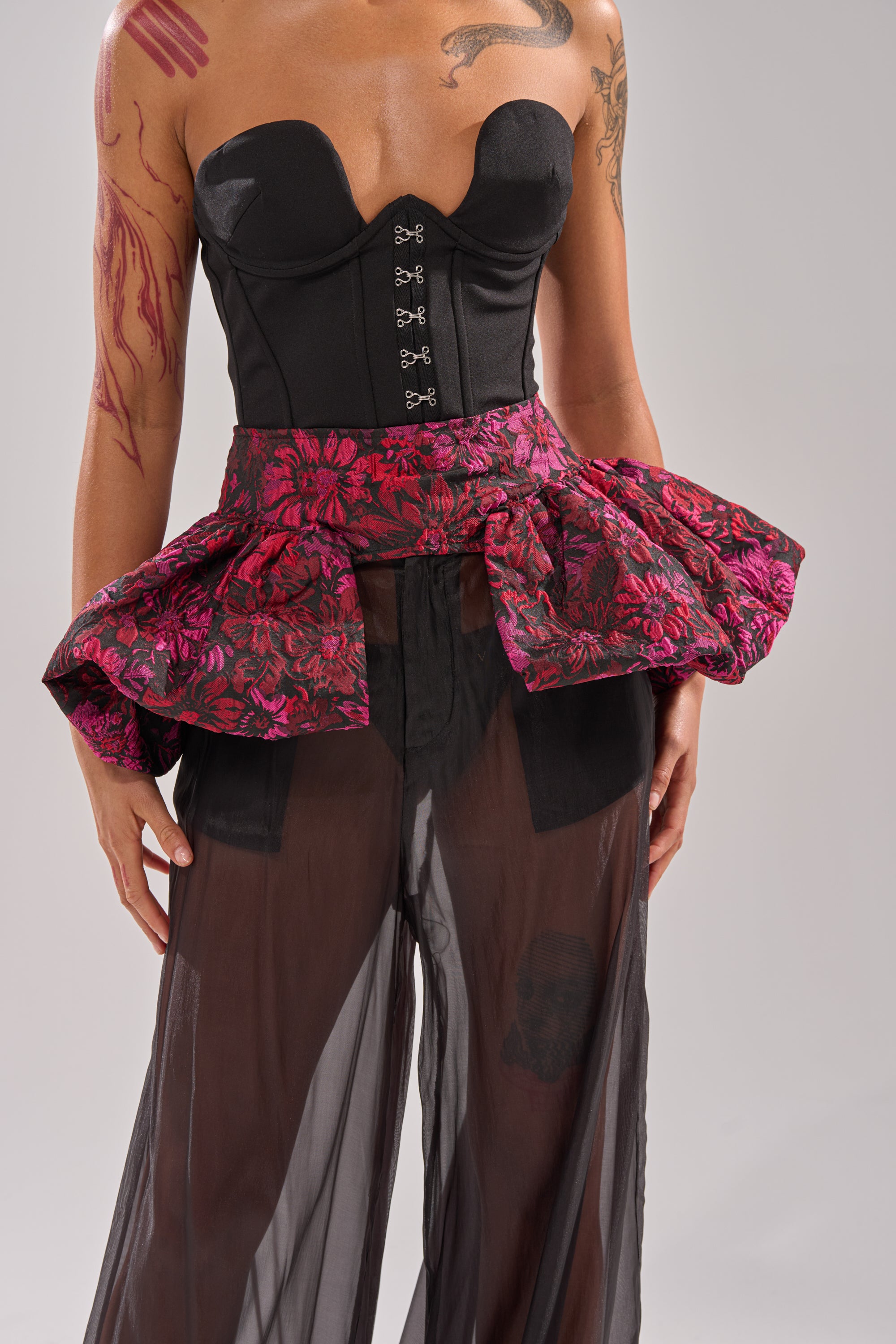 Wearing a black strapless corset with hook fastenings, sheer black pants, and the NIGHTLY FLORAL PEPLUM BELT in red and black jacquard, this look is accented by visible tattoo-like body art for a gothic-chic effect.