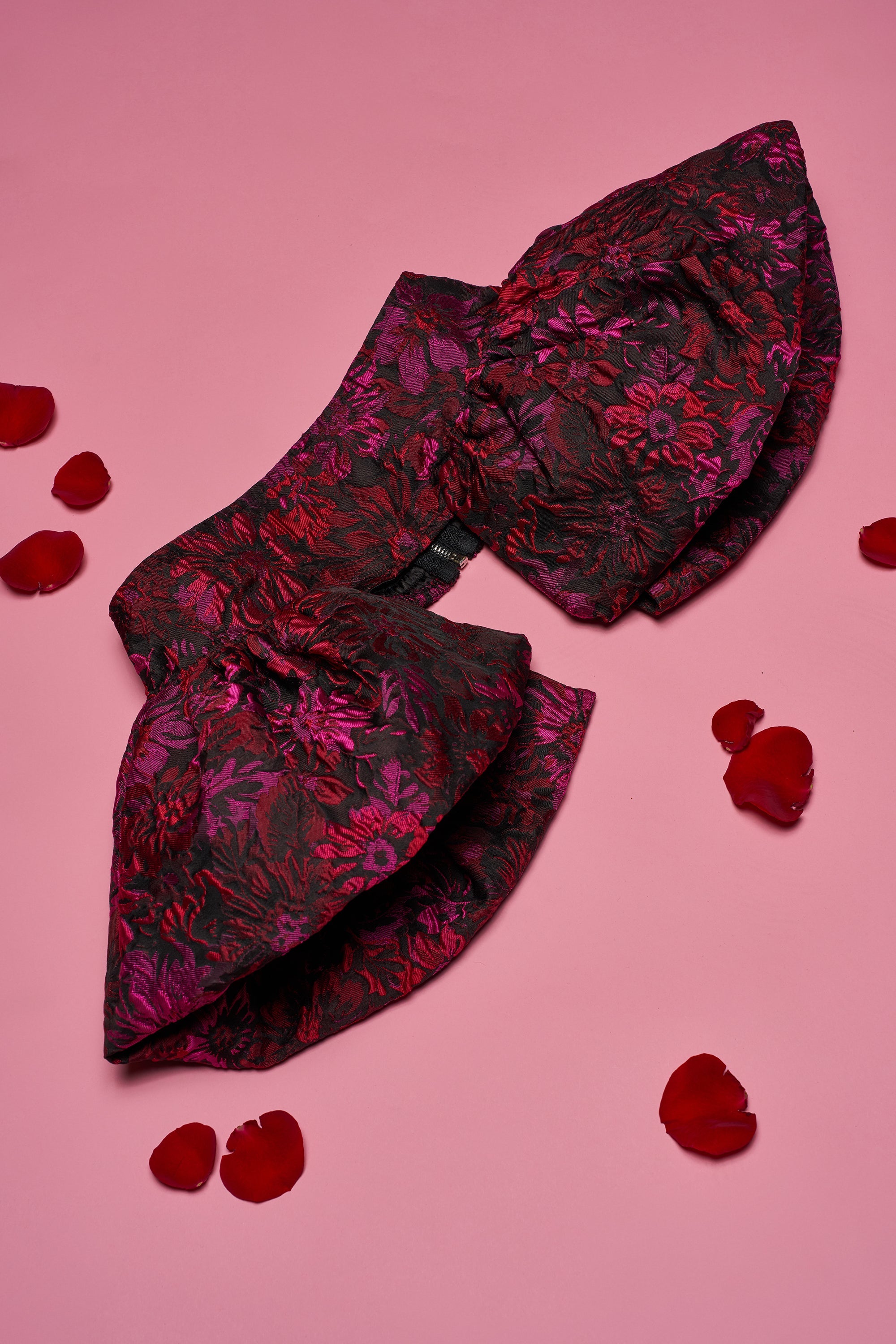 The NIGHTLY FLORAL PEPLUM BELT, featuring a dark floral pattern with red and purple hues inspired by gothic chic trends, is displayed on a pink surface surrounded by scattered red rose petals. 0
