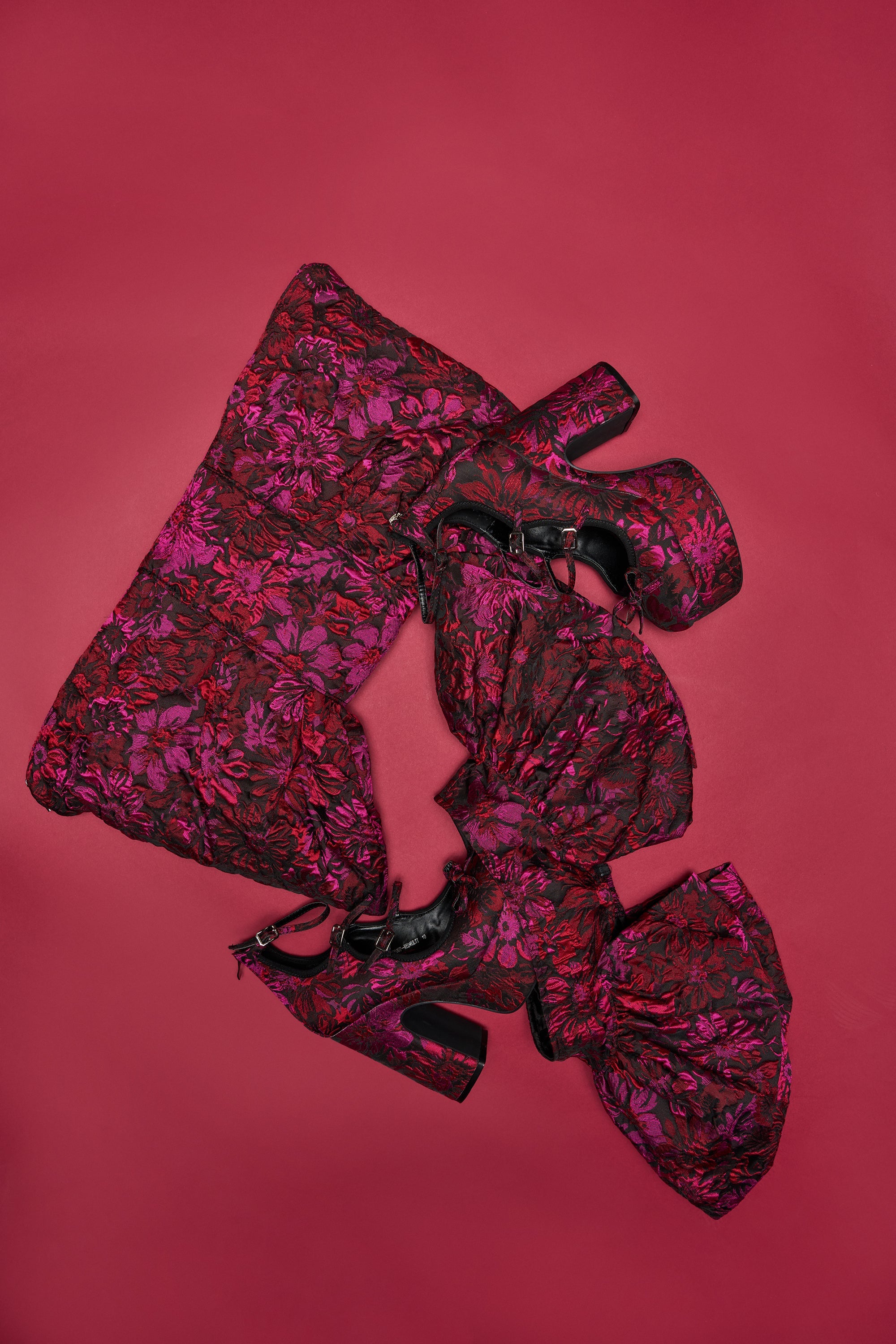 The NIGHTLY FLORAL PEPLUM BELT, featuring a black, pink, and red floral jacquard design with large bows, is displayed on a matching clutch against a solid red background. 1