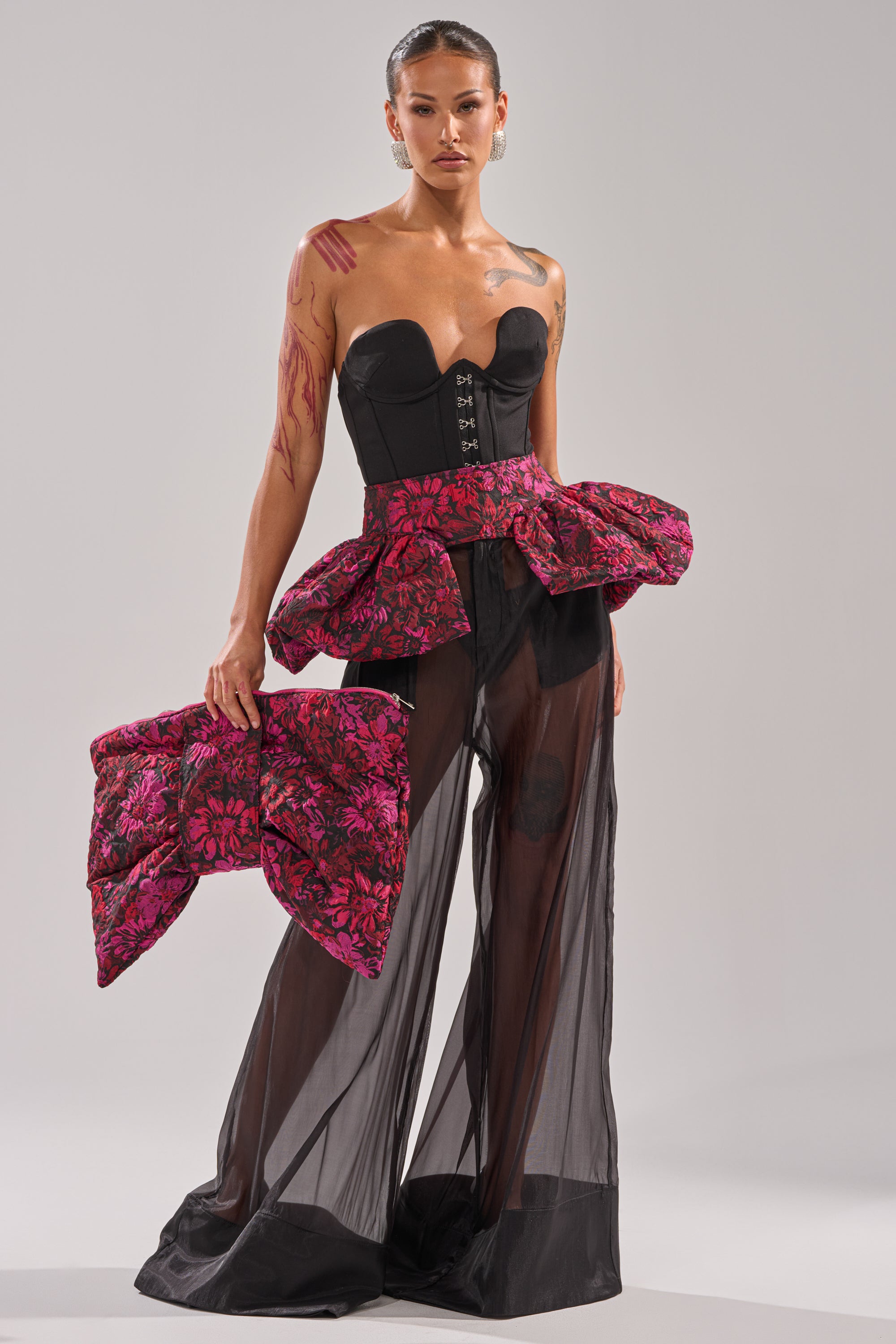 A woman styles the NIGHTLY FLORAL PEPLUM BELT with a black strapless corset, sheer black pants, and a matching oversized bow, accessorized with gothic-chic pieces against a plain light background.