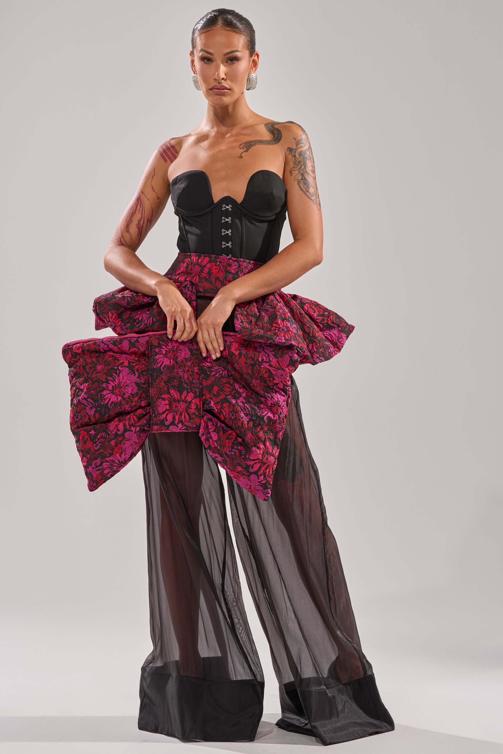 A woman with slicked-back hair and arm tattoos faces forward wearing a black strapless corset, sheer black wide-leg pants, and the NIGHTLY FLORAL PEPLUM BELT—a voluminous pink jacquard skirt accent piece.