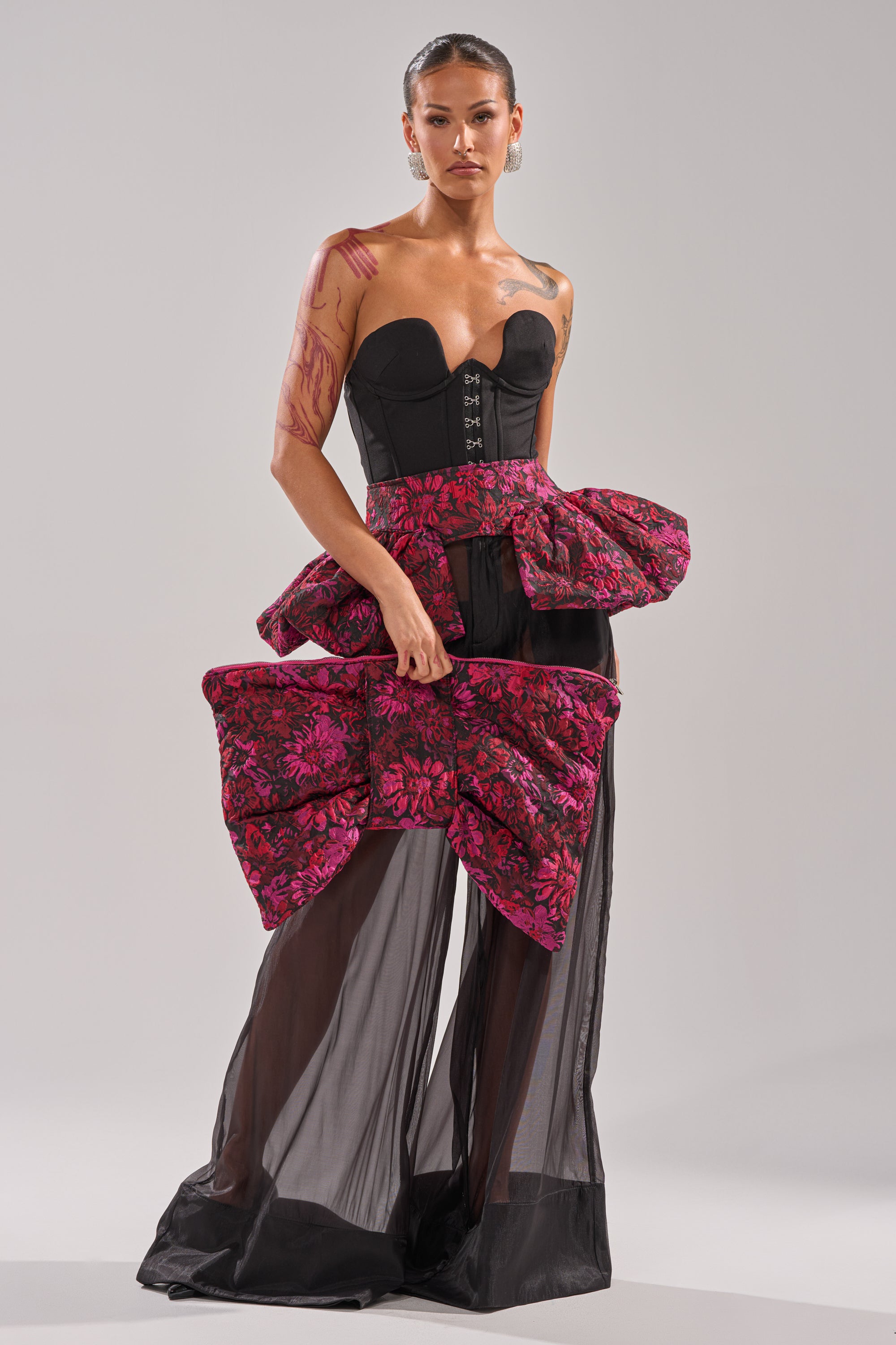 A woman wears the NIGHTLY FLORAL PEPLUM BELT over a strapless black corset top and sheer skirt, accented with large pink bows, jacquard details, and purple body paint for a bold, modern look.