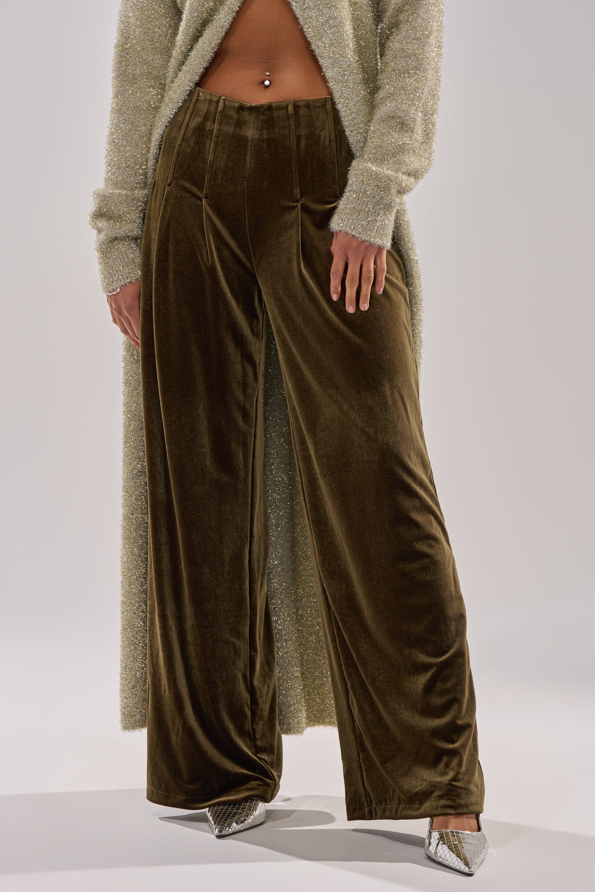 Someone wears SHOW ME IT TROUSER in brown velvet with a high-rise, wide-leg fit, paired with a shimmery light-tone long cardigan and pointed silver heels. The midriff and hands are visible against a plain, light backdrop.