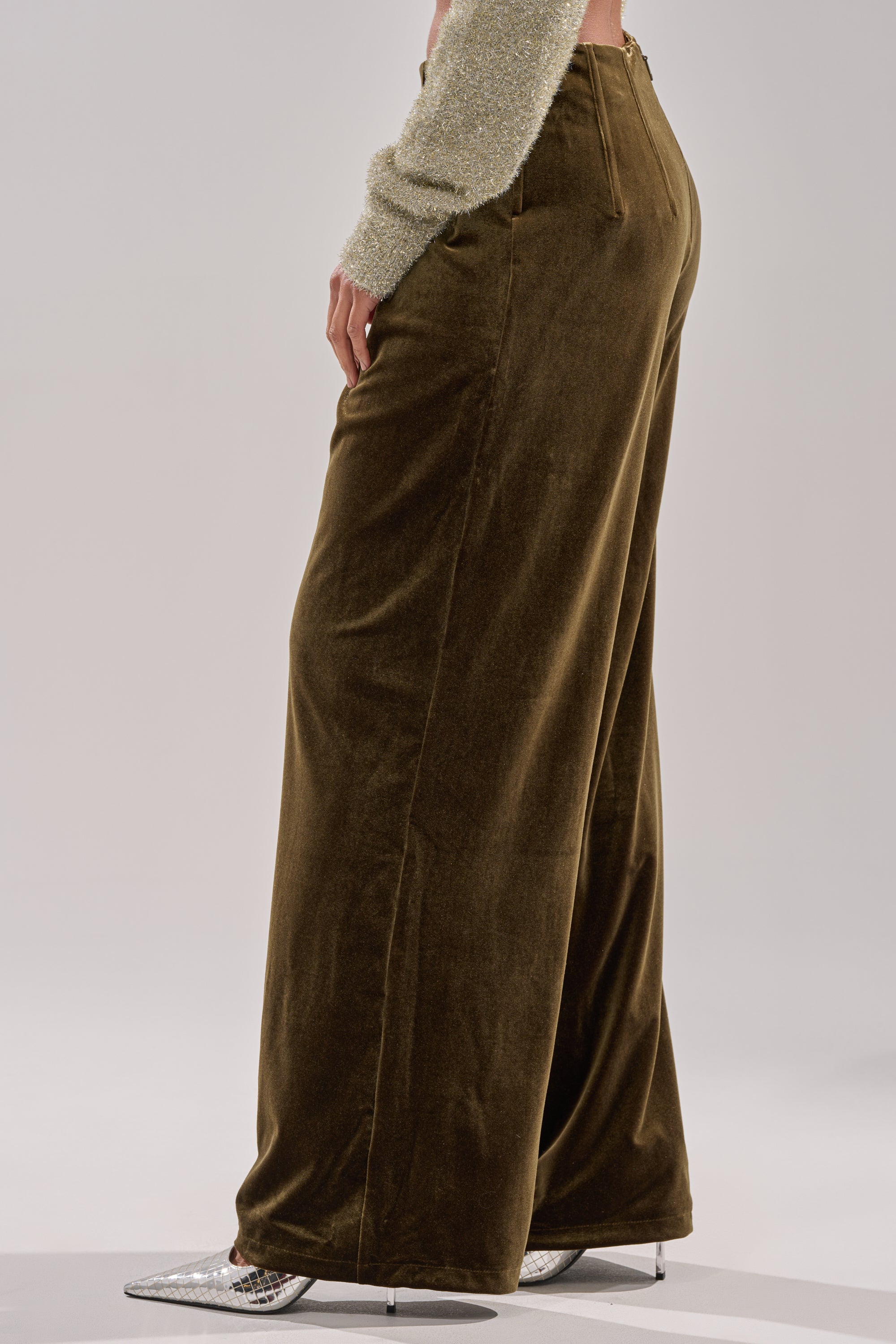 A person wears the SHOW ME IT TROUSER in brown velvet, styled with a sparkly light-colored long-sleeve top and silver pointed-toe heels, shown from the waist down and back view.