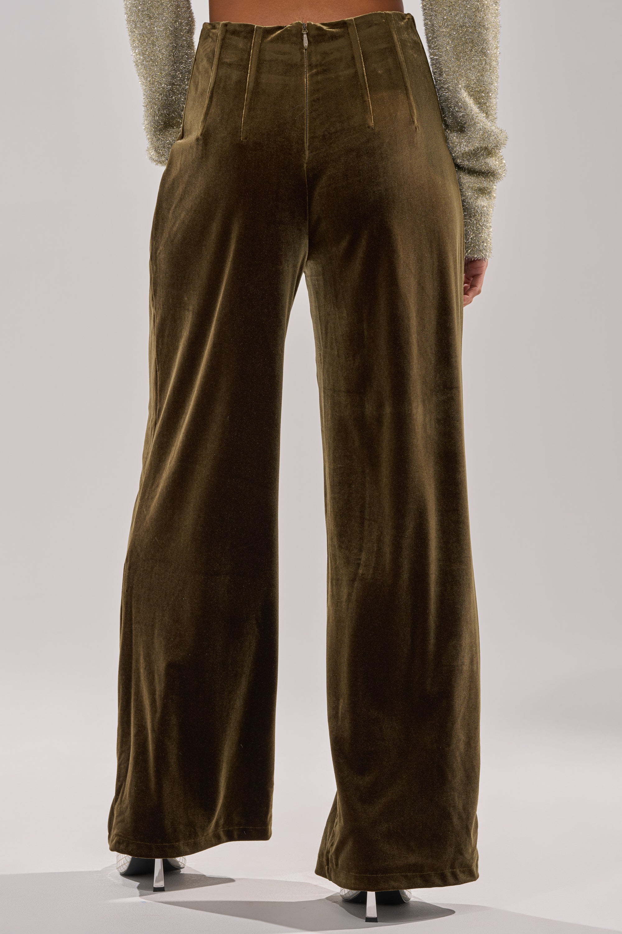 A person shown from the back stands against a plain light background, wearing the SHOW ME IT TROUSER—brown velvet, high-rise fit, wide-leg style—with a sparkly long-sleeve top.