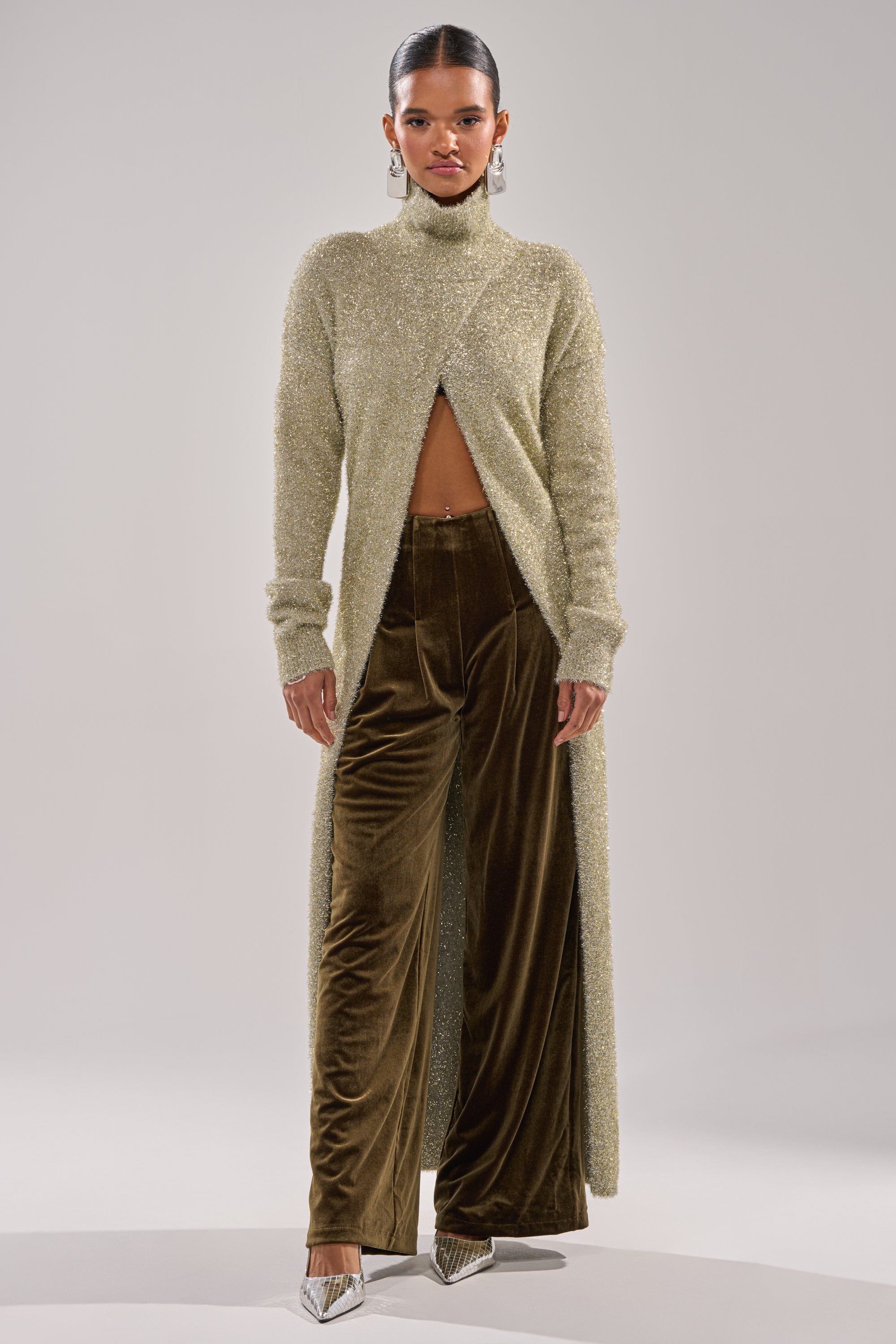 A woman stands against a plain background wearing a sparkly beige long-sleeve turtleneck with a front slit, paired with the SHOW ME IT TROUSER and silver pointed-toe heels, revealing her midriff.
