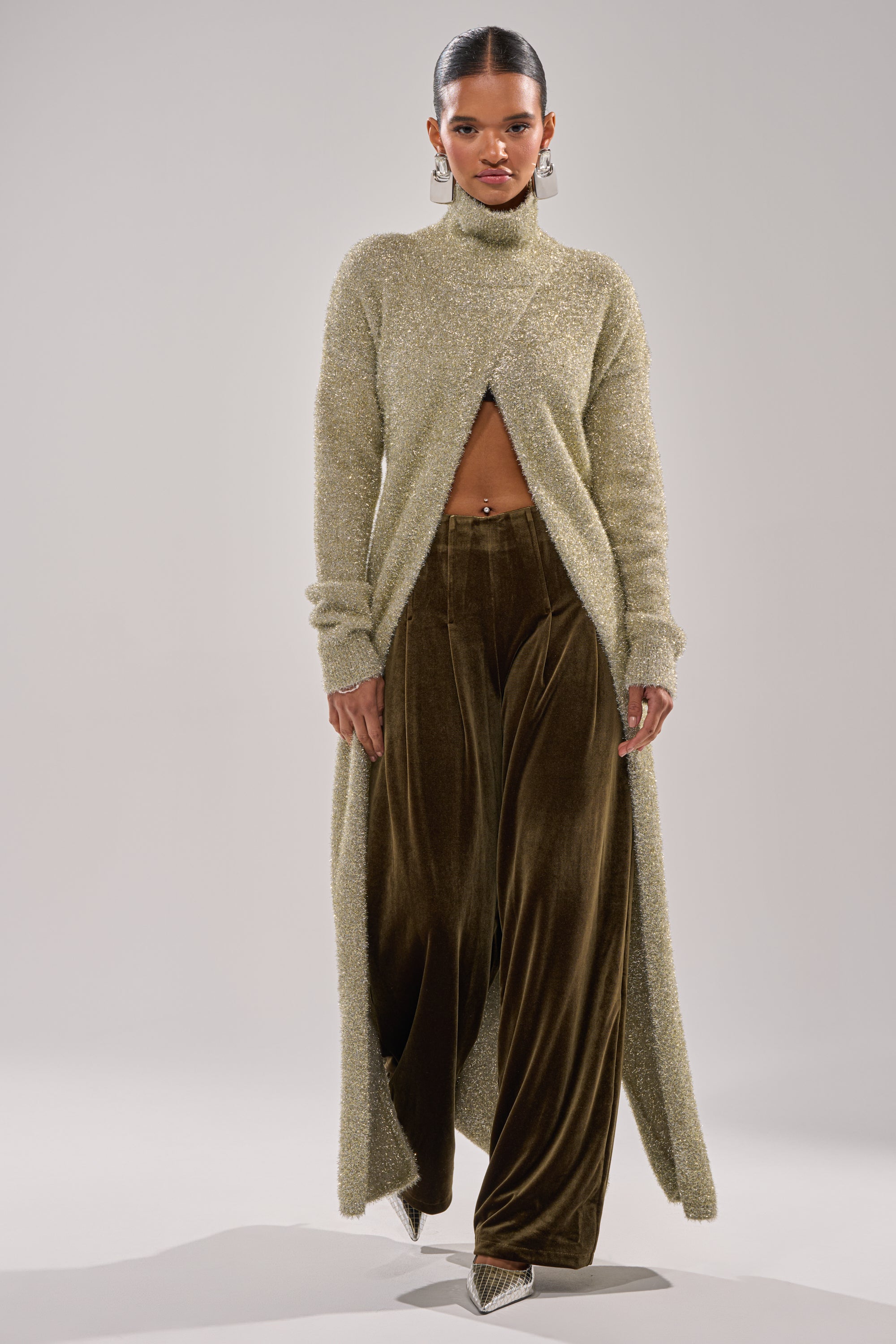 A woman models the SHOW ME IT TROUSER, styled with a long, textured light green turtleneck sweater with a front slit and pointed heels, against a plain light background.