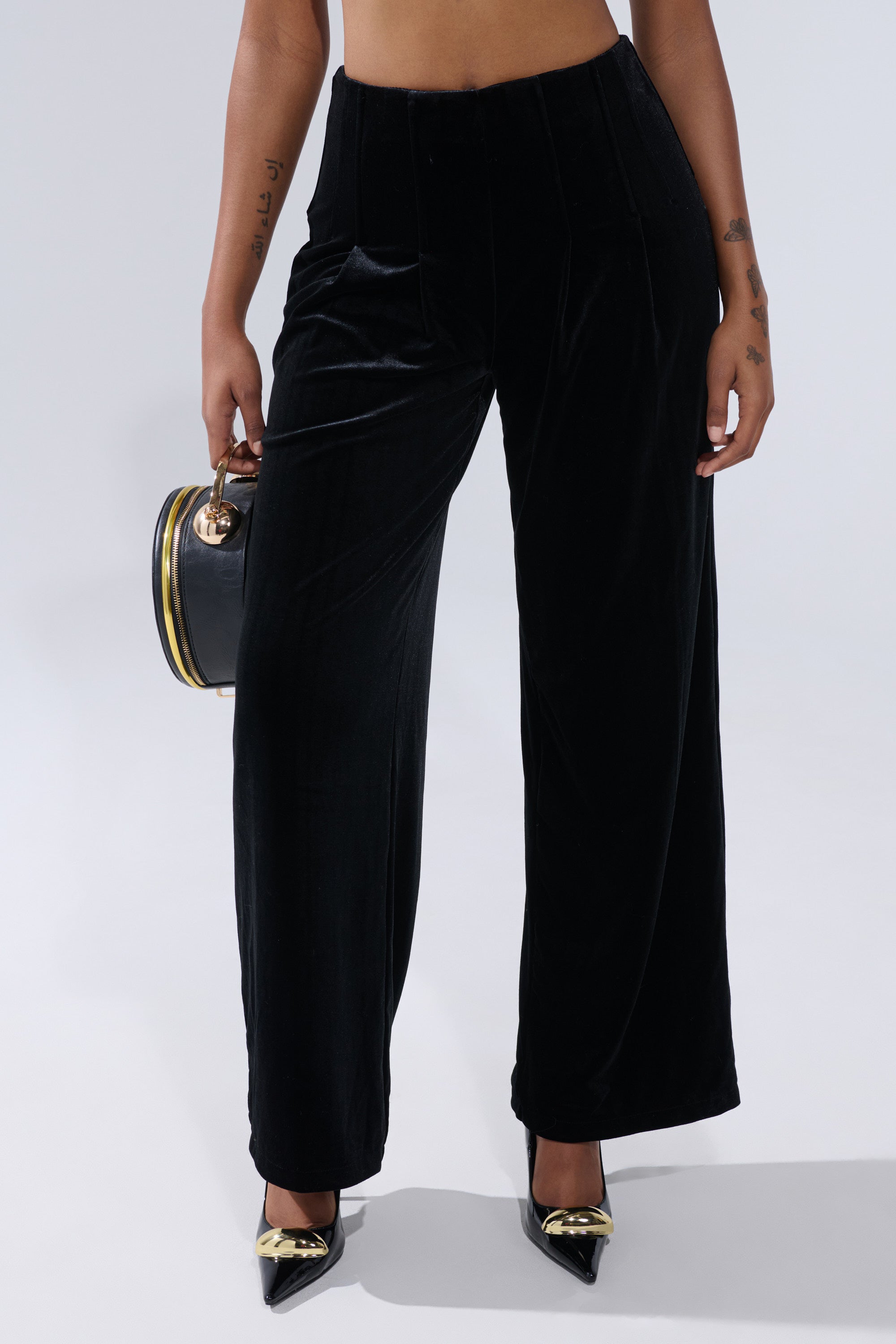 A person wears the SHOW ME IT VELVET TROUSER—high-rise, wide-leg black velvet pants—paired with black pointed-toe heels featuring gold accents and a black handbag with gold details, set against a light gray background. 0
