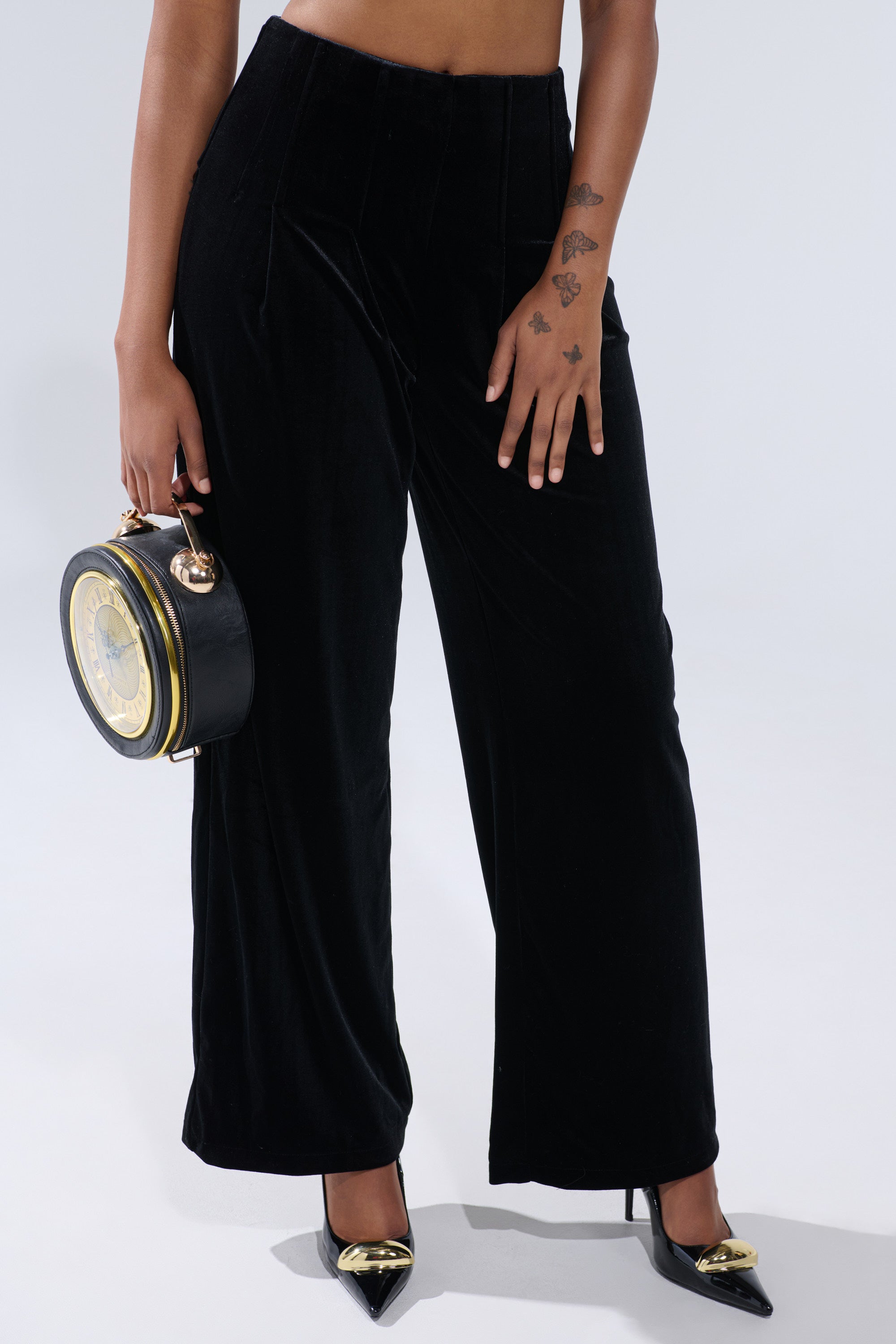 Wearing the SHOW ME IT VELVET TROUSER, a person pairs these high-rise wide-leg black velvet pants with black heels featuring gold accents and holds a round black handbag with a clock face—only their lower half is shown. 1