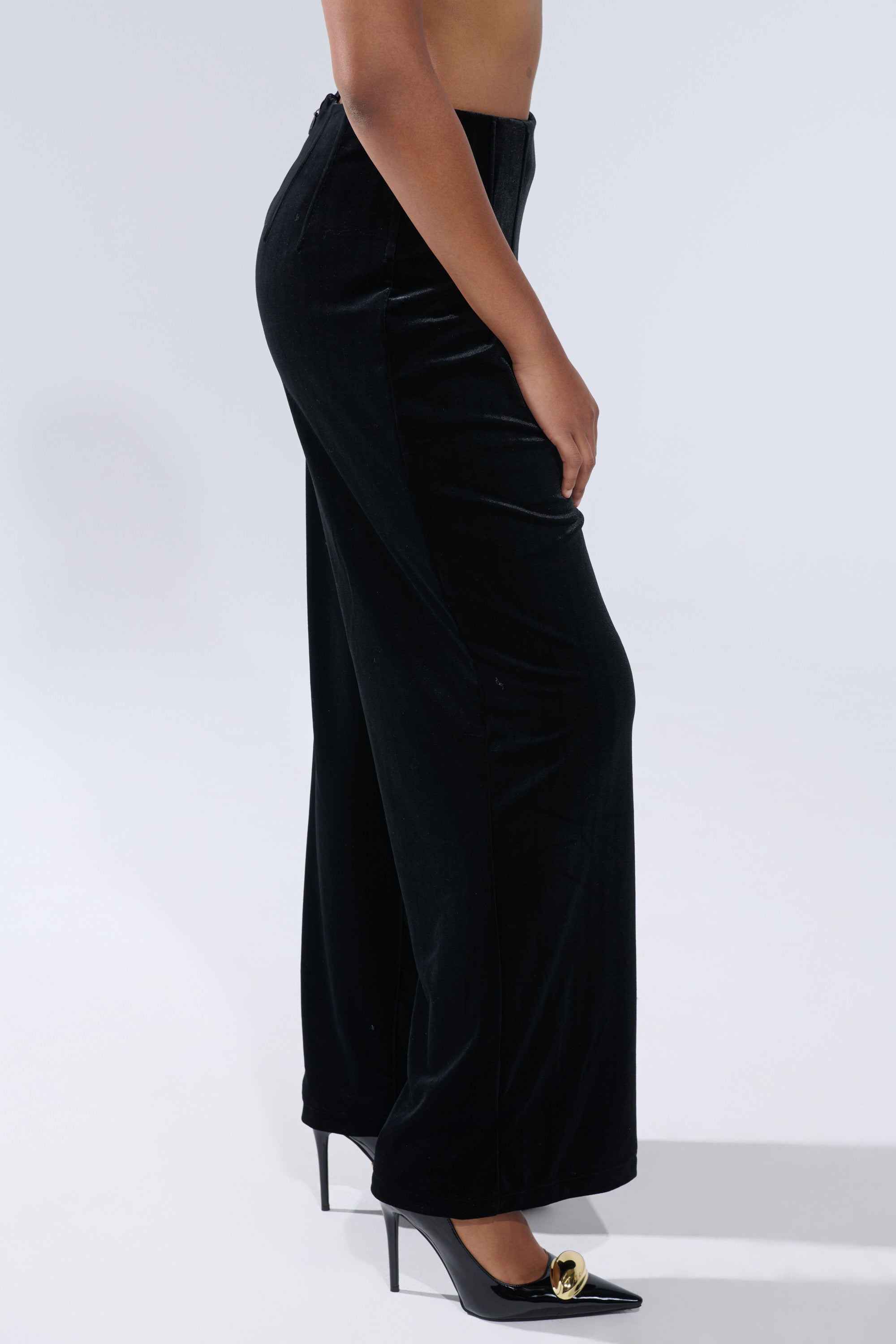 A person stands in profile against a plain light background, wearing the SHOW ME IT VELVET TROUSER—high-rise, wide-leg black velvet pants—paired with black pointed-toe heels. 2
